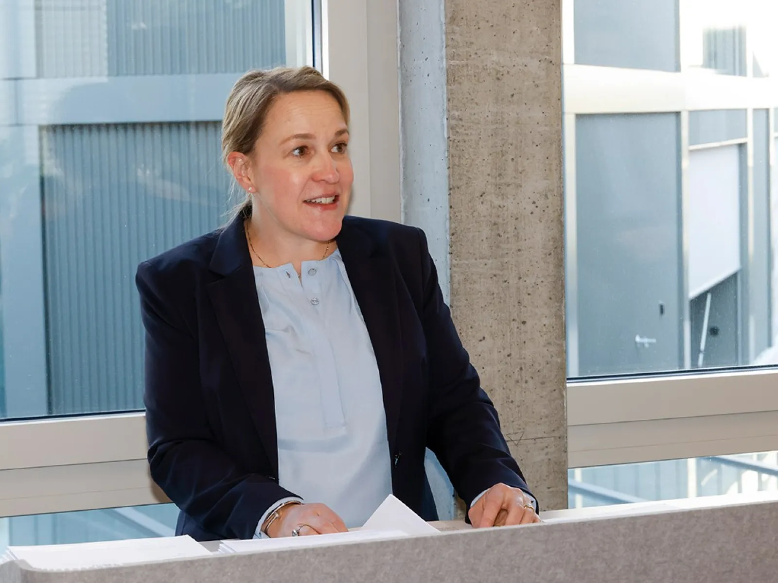 Eine Person hält eine Rede in einem modernen Gebäude. Sie steht hinter einem Rednerpult vor einem großen Fenster.