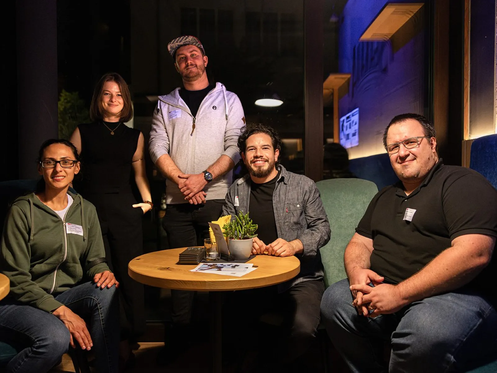 Eine Gruppe von fünf Personen sitzt und steht um einen Tisch in einem gemütlichen Café. Sie lächeln in die Kamera und wirken entspannt. Auf dem Tisch liegen Zeitschriften und eine Pflanze. Die Szene ist warm beleuchtet, was eine einladende Atmosphäre schafft.