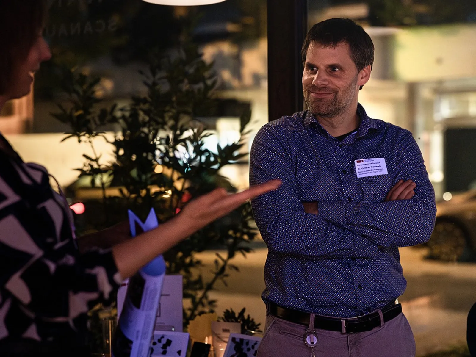 Eine Person steht lächelnd mit verschränkten Armen in einem Raum, während eine andere Person gestikuliert. Im Hintergrund sind Pflanzen und verschwommene Lichter zu sehen, was auf eine entspannte Atmosphäre bei einem Alumni-Treffen der Universität Liechtenstein hindeutet.