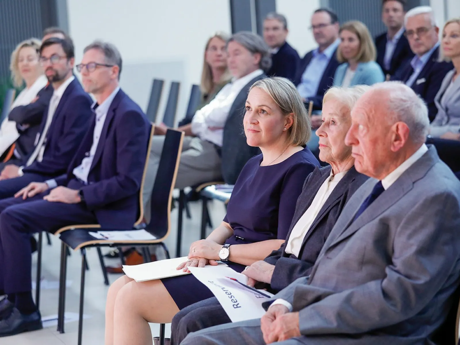 Eine Gruppe von Menschen sitzt in einem Auditorium. Im Vordergrund sitzen drei Personen in der ersten Reihe, aufmerksam nach vorne blickend. Die Veranstaltung scheint formell zu sein, mit mehreren Reihen von Teilnehmern im Hintergrund.