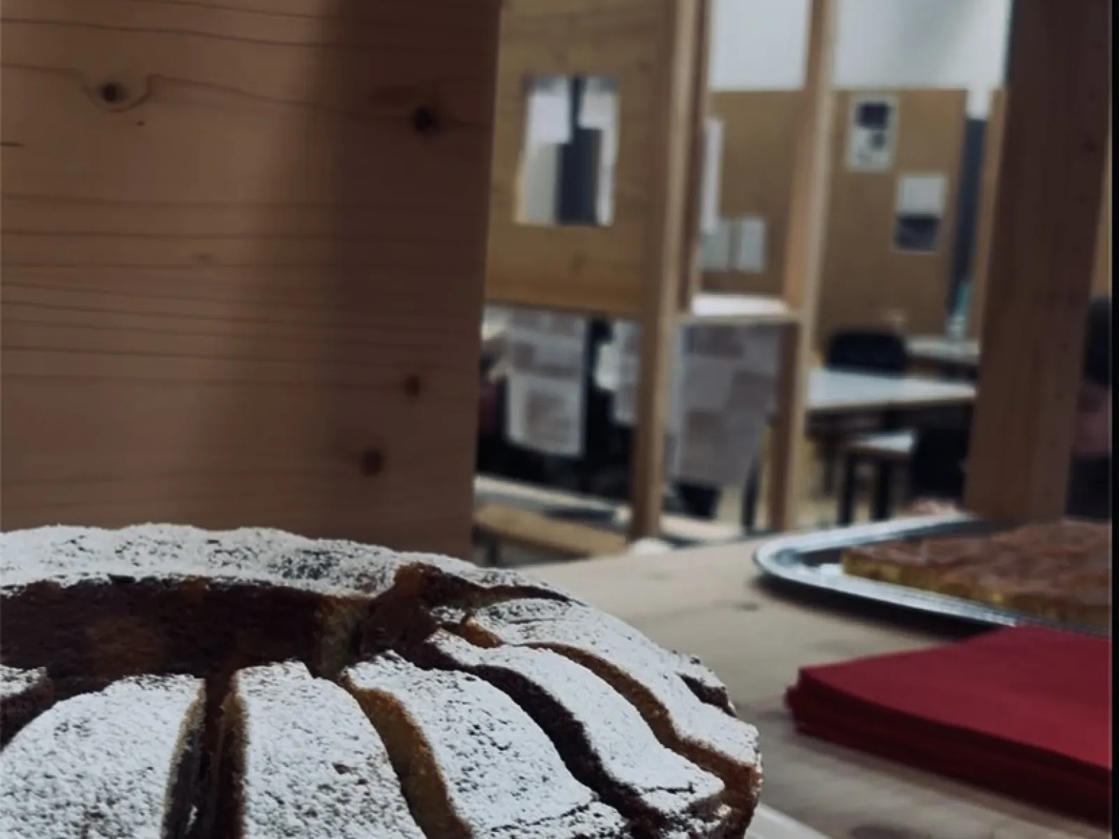 Ein frisch gebackener, mit Puderzucker bestäubter Kuchen steht auf einem Holztisch. Im Hintergrund sind Holzkonstruktionen und Tische in einem Raum zu sehen, die eine kreative und einladende Atmosphäre vermitteln.