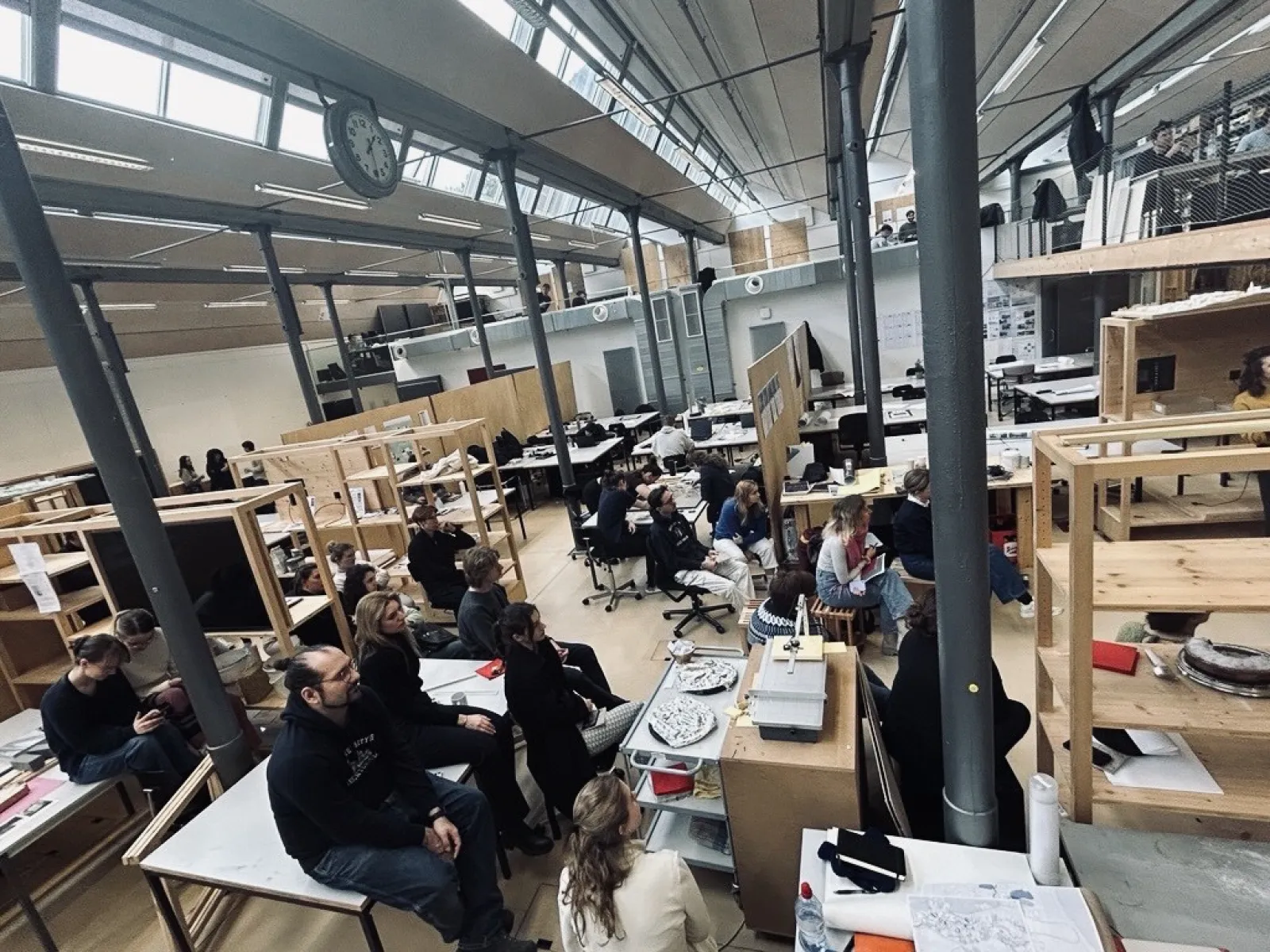 Ein großer, heller Raum mit hohen Decken in der Liechtenstein School of Architecture. Studierende sitzen an Tischen und hören einer Präsentation zu. Im Vordergrund sind Architekturmodelle und Zeichnungen zu sehen. Die Atmosphäre wirkt konzentriert und kreativ.