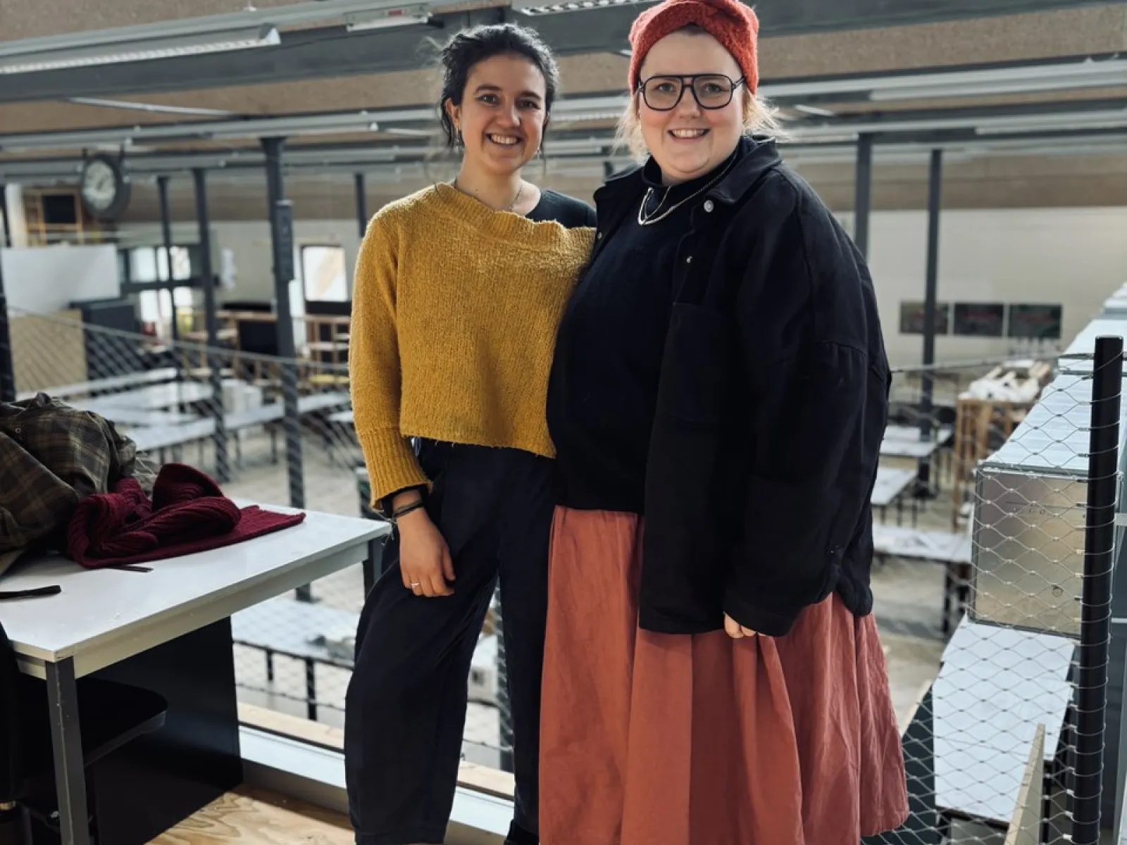 Zwei Personen stehen lächelnd in einem modernen Architekturatelier mit großen Fenstern und industriellem Design. Sie tragen lässige, kreative Kleidung. Im Hintergrund sind Arbeitsplätze und eine Uhr sichtbar.