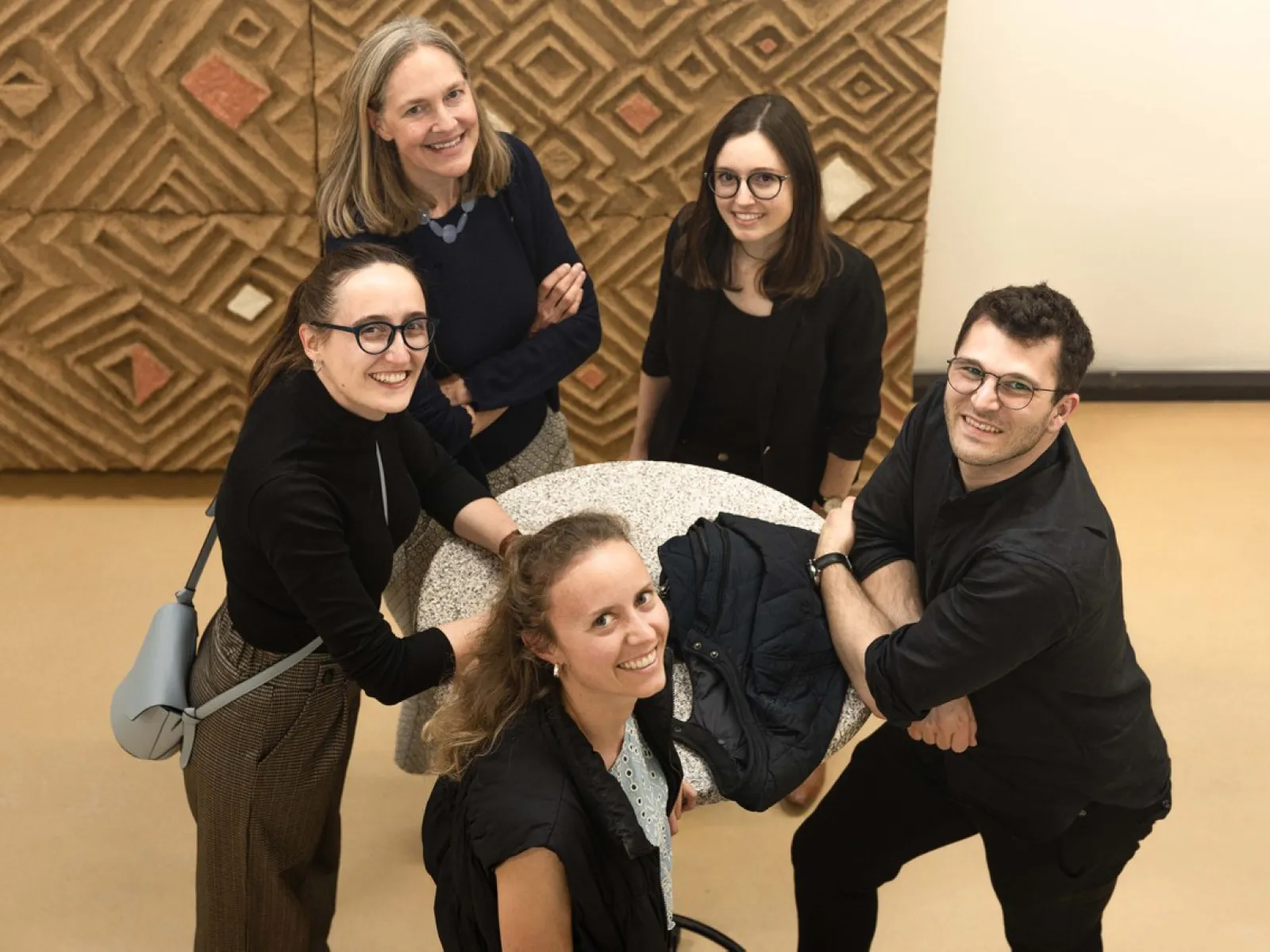 Eine Gruppe von fünf Personen steht um einen runden Tisch und lächelt in die Kamera. Der Hintergrund zeigt ein geometrisches Muster an der Wand. Die Szene wirkt freundlich und einladend.