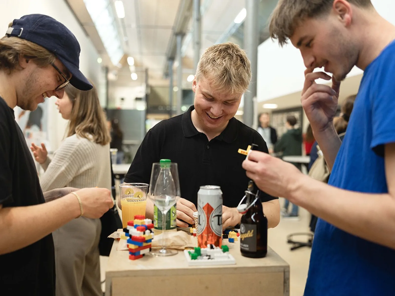 Drei junge Erwachsene stehen um einen Tisch mit Getränken und Snacks, lachen und interagieren miteinander. Im Hintergrund sind weitere Personen in einer hellen, modernen Umgebung zu sehen.