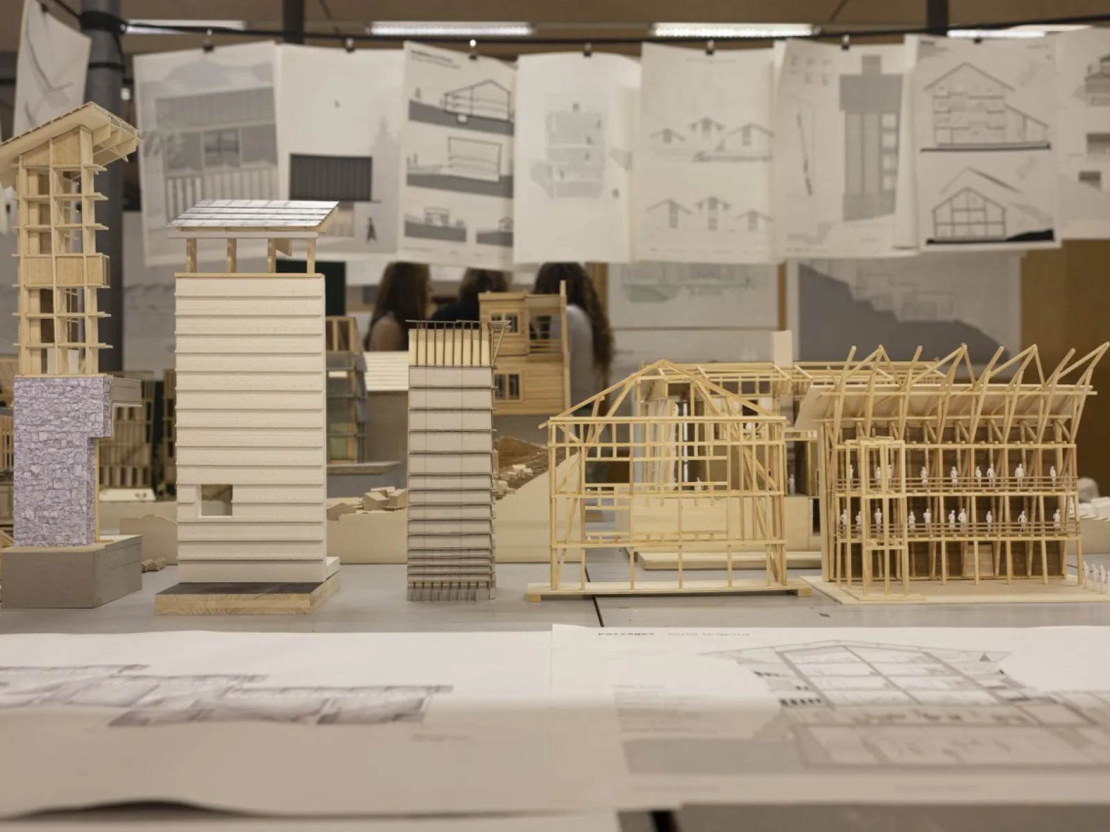 Architekturmodelle aus Holz und Papier stehen auf einem Tisch, umgeben von aufgehängten Architekturzeichnungen. Im Hintergrund sind unscharf Personen zu erkennen.