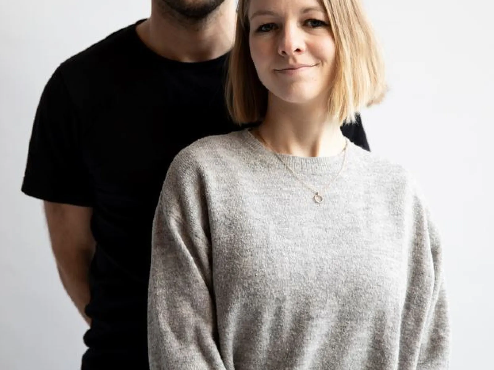 Ein Mann und eine Frau stehen vor einem neutralen Hintergrund. Beide blicken direkt in die Kamera. Die Frau trägt einen grauen Pullover, der Mann ein schwarzes T-Shirt.