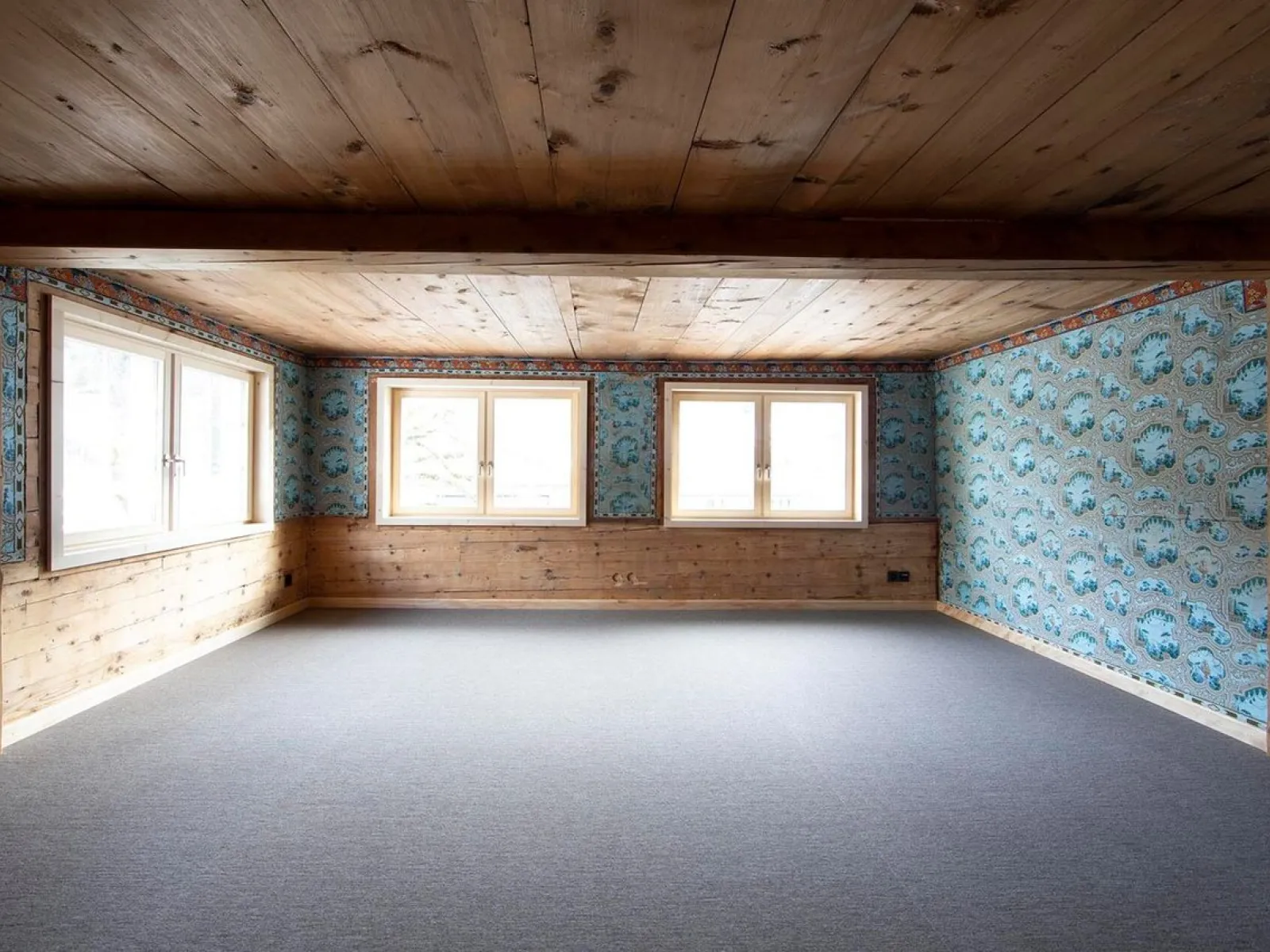 Ein leerer Raum mit Holzbalkendecke und Holzwänden, teilweise mit blauer, gemusterter Tapete. Drei große Fenster lassen Tageslicht herein. Der Boden ist mit grauem Teppich ausgelegt.