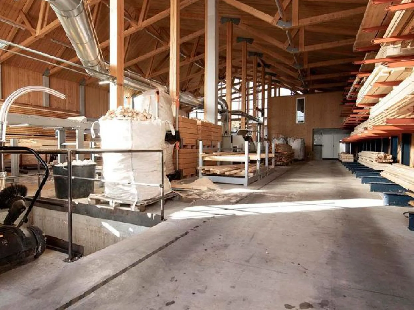 Innenansicht einer modernen Holzwerkstatt mit sichtbaren Holzbalken und gestapelten Holzplanken. Die Werkstatt ist hell erleuchtet und zeigt eine aufgeräumte, funktionale Arbeitsumgebung.