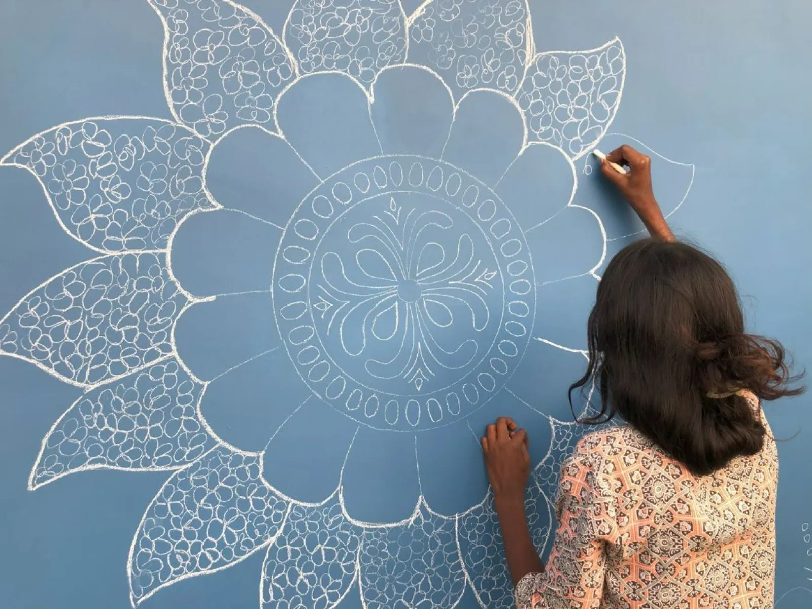 Eine Person zeichnet ein detailliertes, weißes Mandala-Muster auf eine blaue Wand.