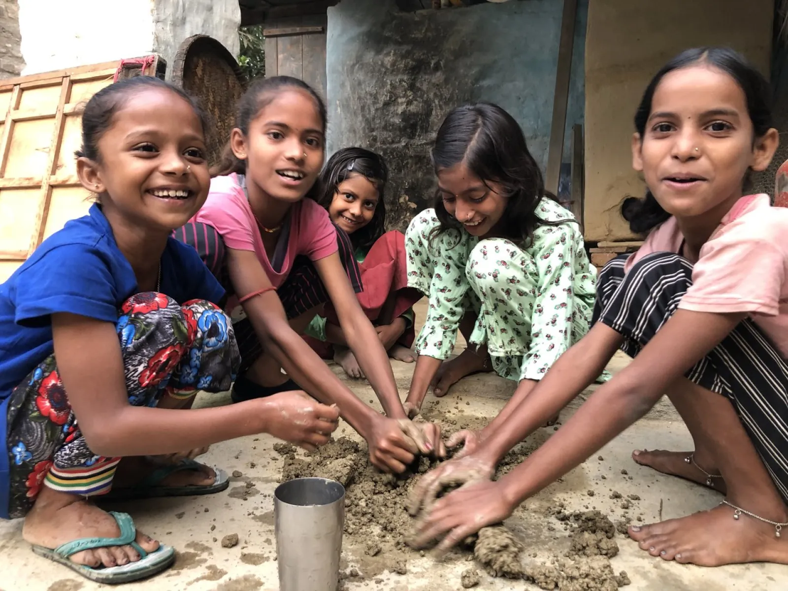 Eine Gruppe von lachenden Kindern kniet auf dem Boden und formt gemeinsam Ton oder Lehm. Die Szene vermittelt Freude und Zusammenarbeit in einem handwerklichen Kontext.