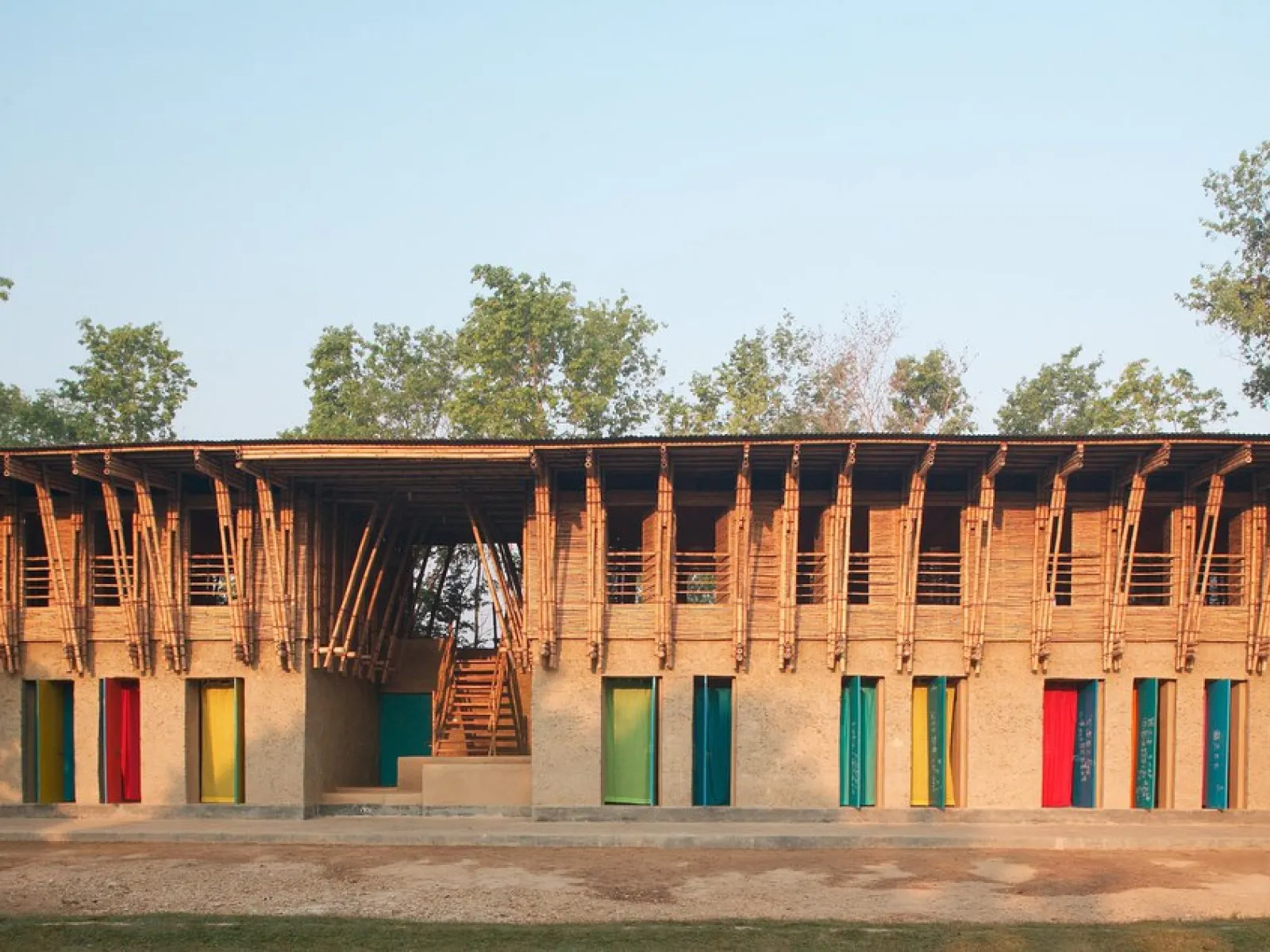 Ein zweistöckiges Gebäude aus natürlichen Materialien wie Lehm und Bambus. Die Fassade zeigt bunte Türen und Fenster, umgeben von Bäumen. Das Design kombiniert traditionelle Bauweisen mit modernem Stil.