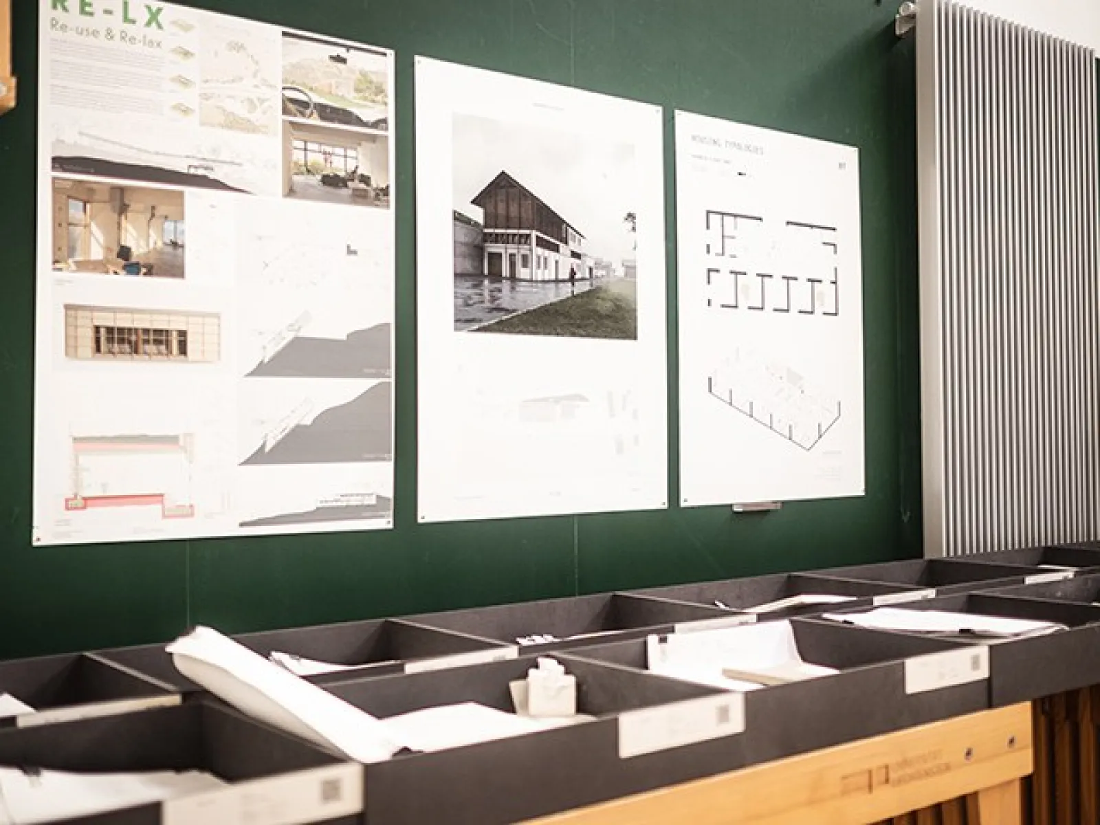 Architektur-Entwürfe und Pläne an einer Wand in der Liechtenstein School of Architecture. Modelle und Zeichnungen sind in Regalen darunter angeordnet.
