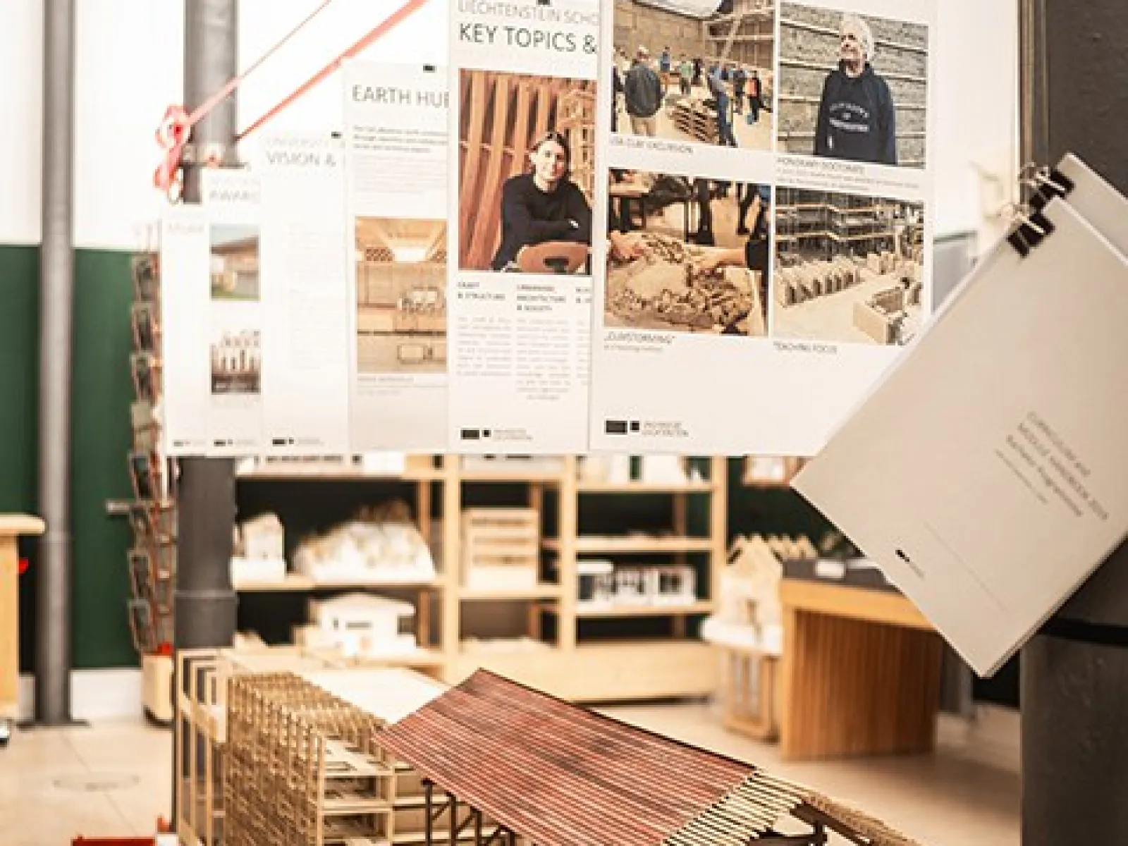 Ein Poster der Liechtenstein School of Architecture zeigt die "Key Topics & Units" mit einem Bild von zwei Personen vor architektonischen Modellen. Im Hintergrund sind weitere Poster zu sehen, die verschiedene Architekturthemen präsentieren.