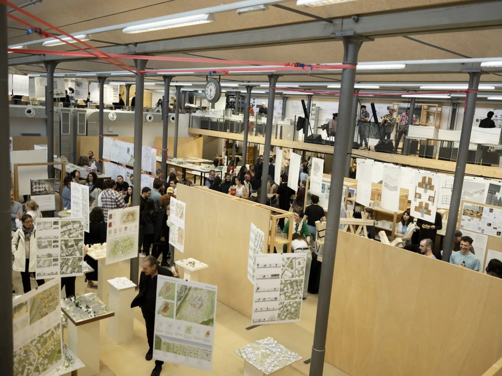 Eine große, gut besuchte Ausstellungshalle mit zahlreichen Architekturmodellen und Plänen, die an Holzwänden präsentiert werden. Menschen bewegen sich durch den Raum, betrachten die ausgestellten Werke und diskutieren. Die Atmosphäre ist lebhaft und interessiert.