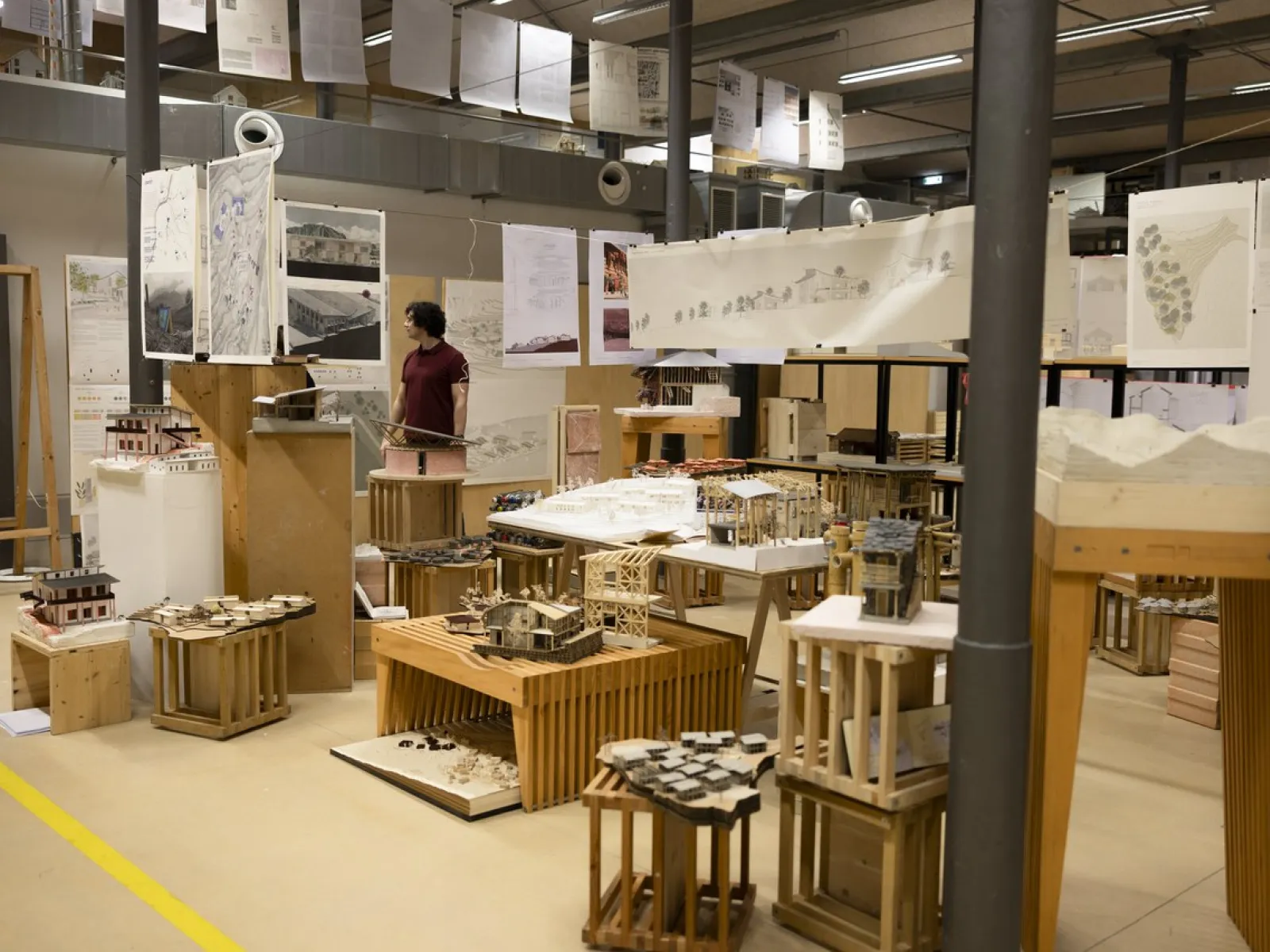 Eine Ausstellung in einem Raum mit Architekturmodellen und Plänen. Eine Person betrachtet die ausgestellten Werke. Die Umgebung wirkt industriell und kreativ, mit zahlreichen Zeichnungen und Modellen auf Tischen und Podesten.