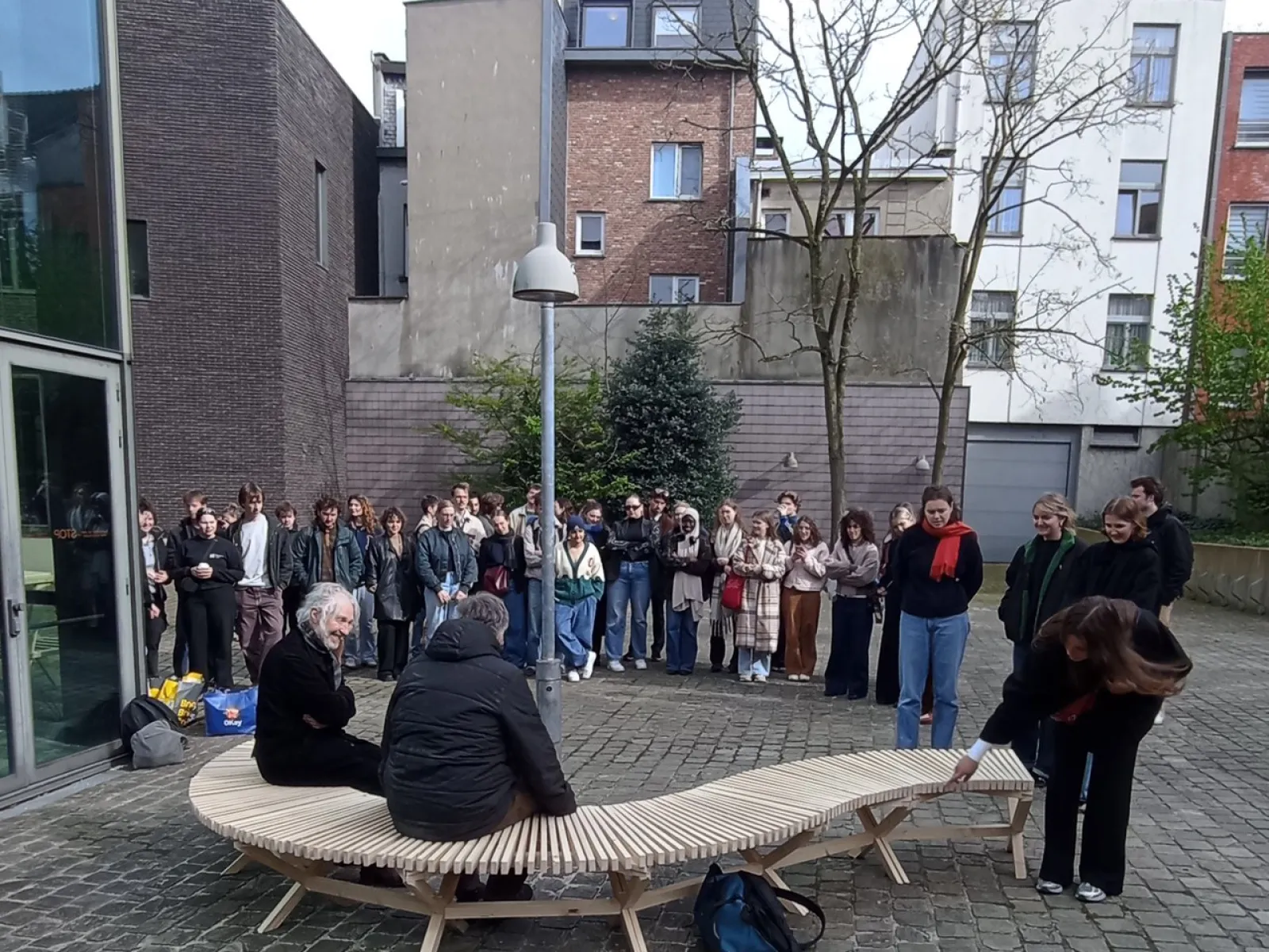 Eine Gruppe von Studierenden steht im Freien um eine geschwungene Holzbank, die von zwei Personen genutzt wird. Die Szene findet auf einem gepflasterten Platz vor modernen Gebäuden statt.