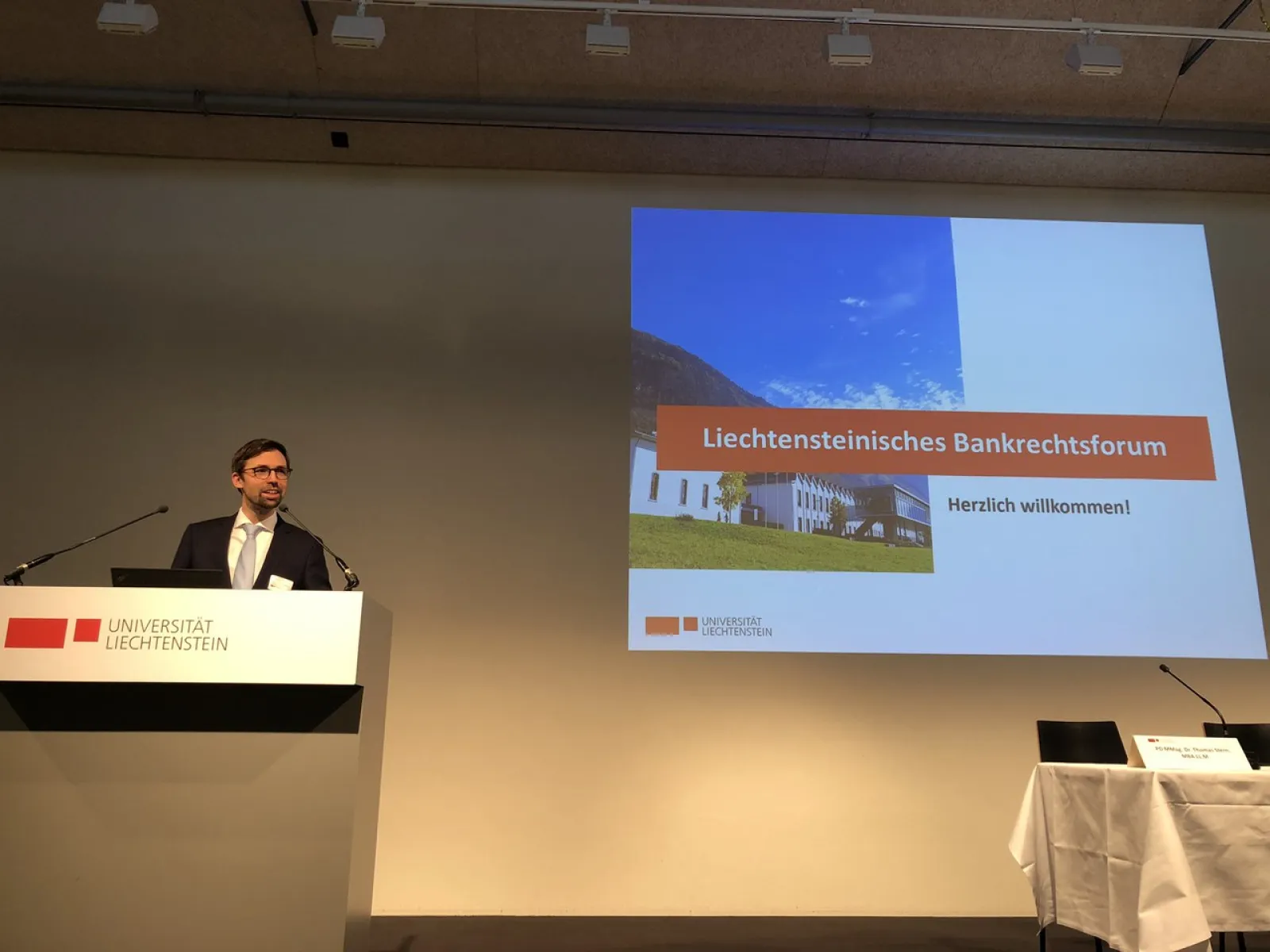 Ein Redner steht am Podium der Universität Liechtenstein während des Liechtensteinischen Bankrechtsforums. Auf der Leinwand ist eine Präsentationsfolie mit der Aufschrift „Liechtensteinisches Bankrechtsforum – Herzlich willkommen!“ zu sehen.