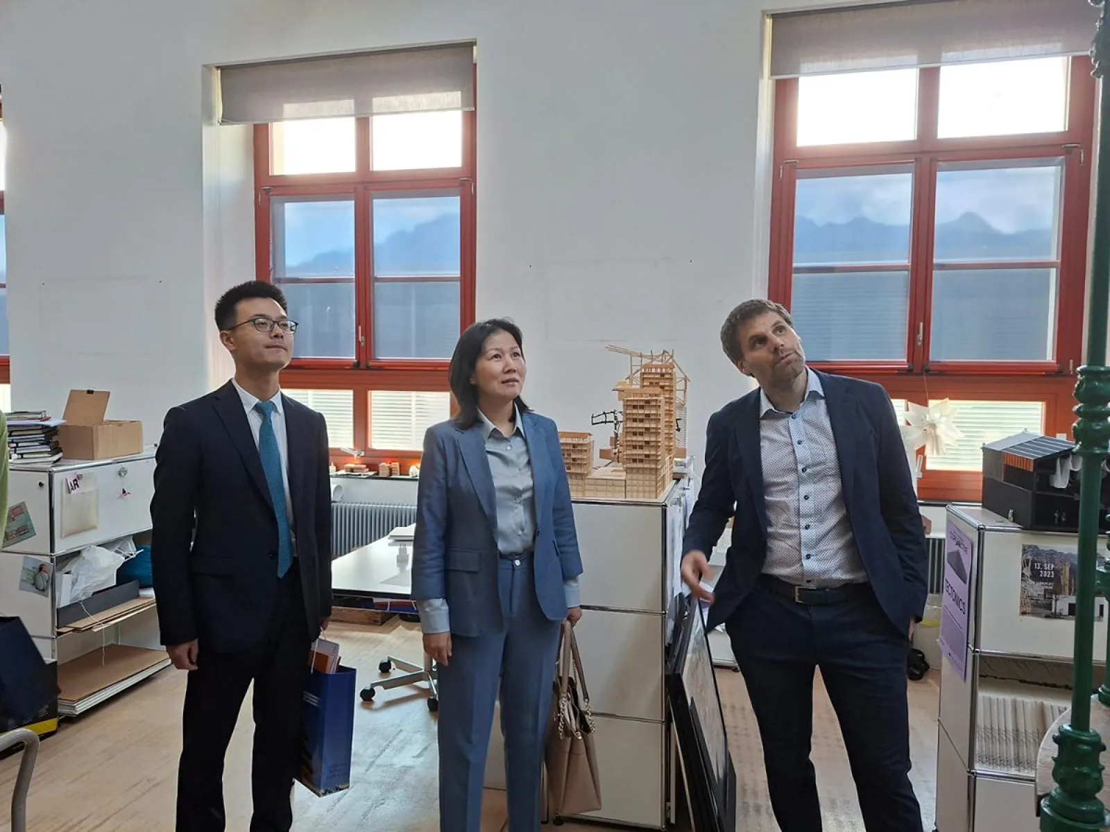 Drei Personen stehen in einem Büro mit großen Fenstern, durch die Berge sichtbar sind. Sie betrachten interessiert etwas außerhalb des Bildes. Im Hintergrund sind Regale und Modelle zu sehen.