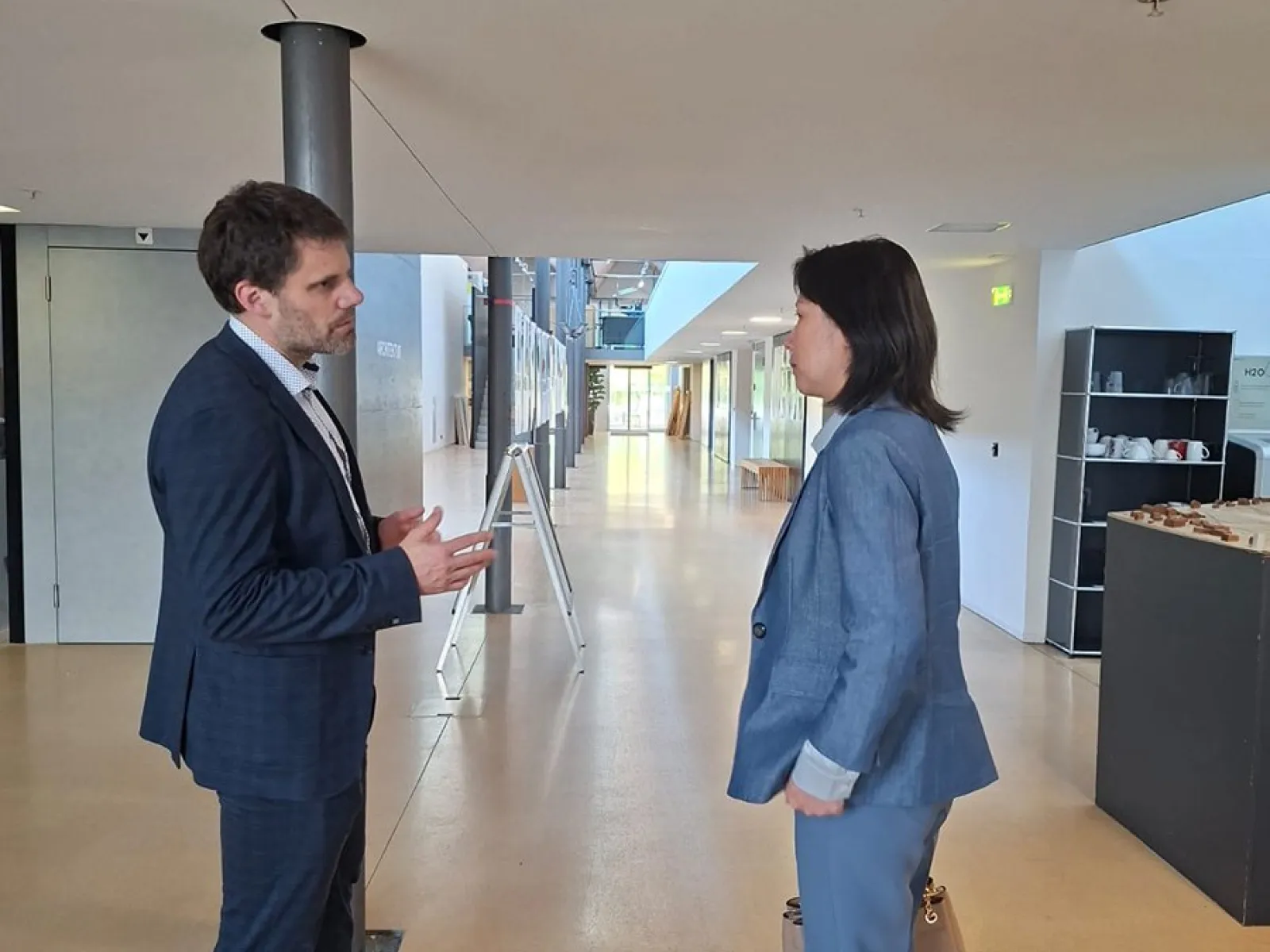 Zwei Personen in einem modernen Gebäude im Gespräch. Beide tragen Business-Kleidung. Im Hintergrund sind helle Flure und moderne Einrichtungselemente zu sehen.