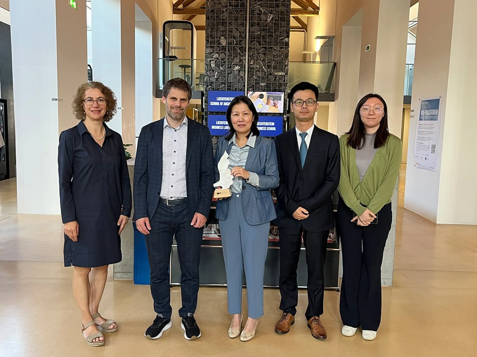 Eine Gruppe von fünf Personen steht in einem modernen Gebäude der Universität Liechtenstein. Sie lächeln in die Kamera. Im Hintergrund sind Informationsschilder der Universität zu sehen.