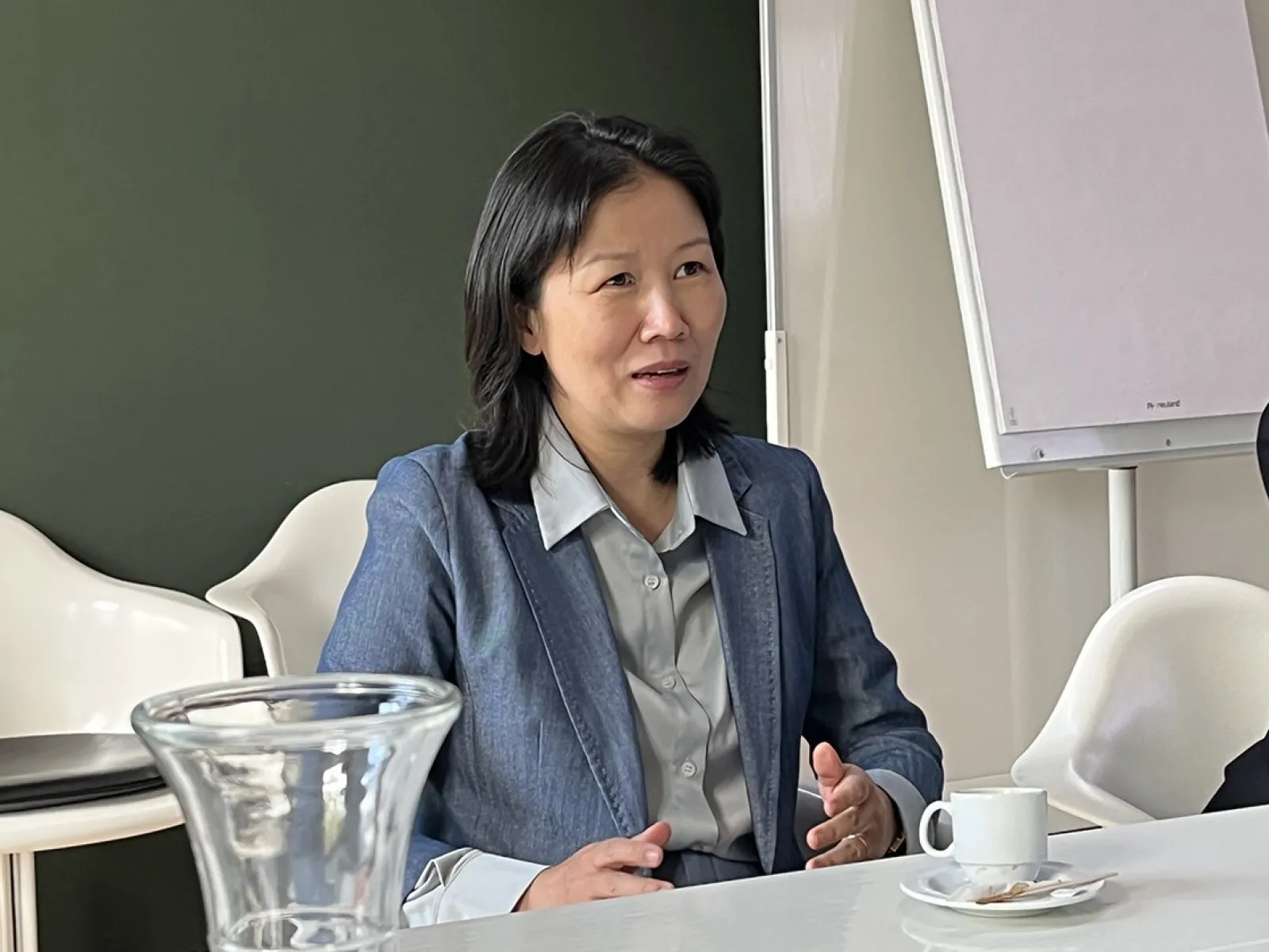 Eine Person in einem blauen Blazer sitzt an einem Konferenztisch mit einer Tasse Kaffee und einem Notizblock. Im Hintergrund ist ein Flipchart zu sehen.