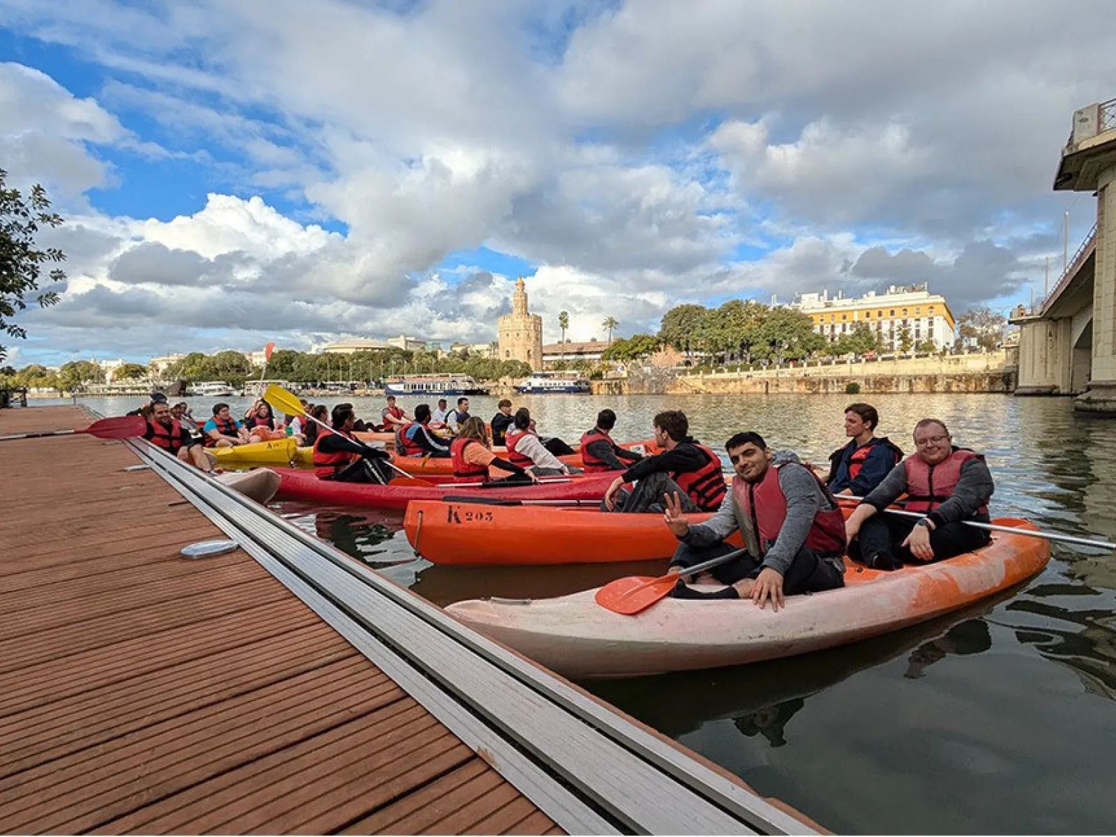 Studierende in Kanus auf einem Fluss in Sevilla, mit Schwimmwesten, vor einer malerischen Stadtlandschaft und bewölktem Himmel.