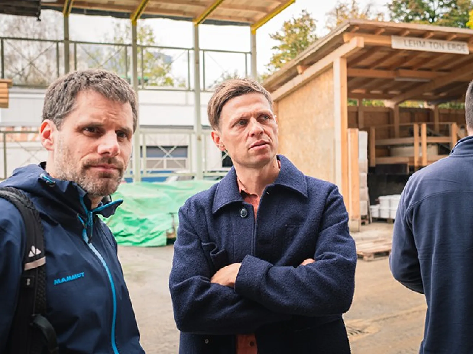 Zwei Männer stehen auf einer Baustelle, umgeben von Holzstrukturen und Baumaterialien. Sie tragen Jacken und schauen aufmerksam in die Ferne. Im Hintergrund sind Baugerüste und ein überdachter Bereich zu sehen.