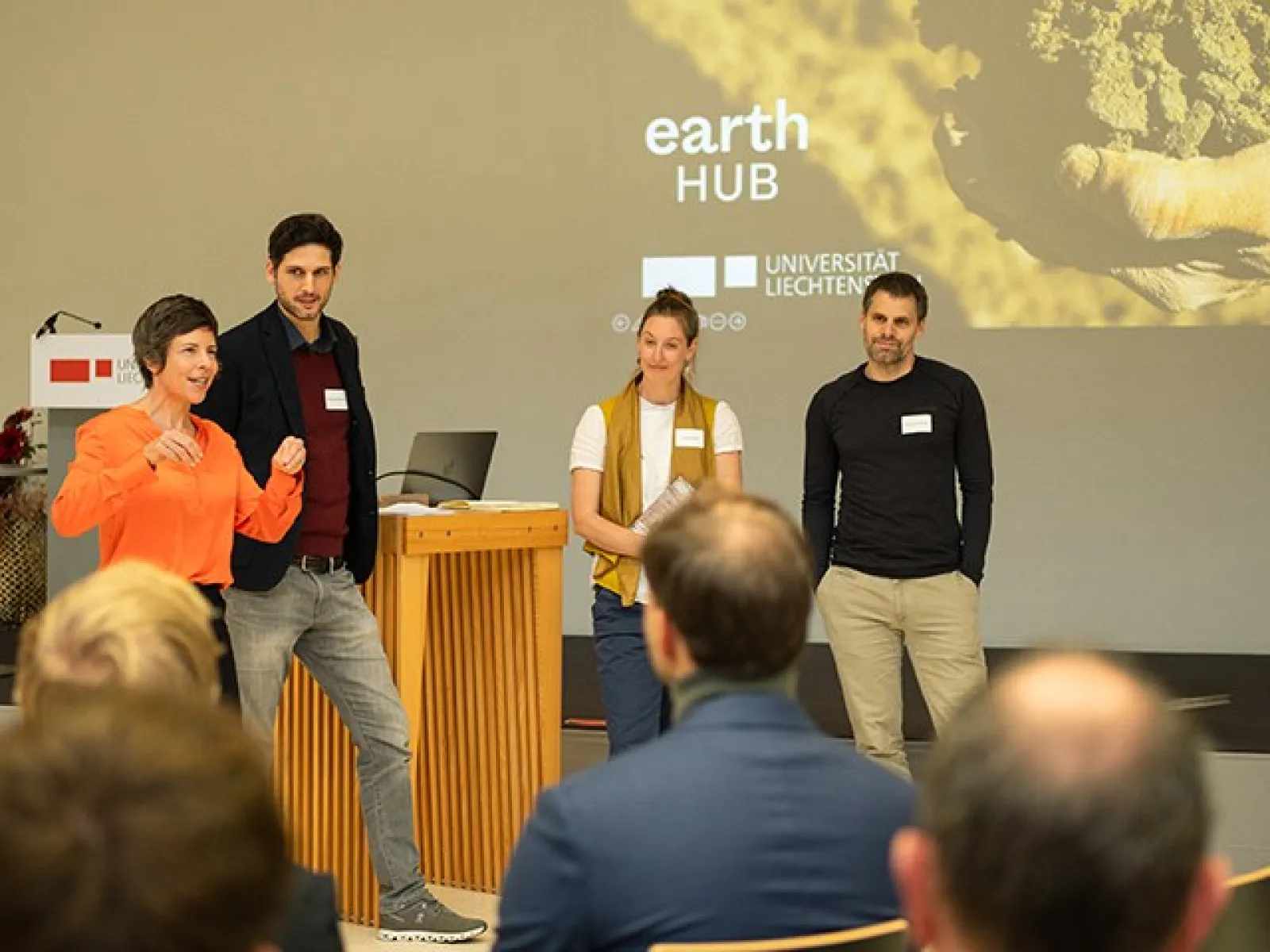 Eine Gruppe von vier Personen steht vor einem Publikum und spricht auf einer Bühne. Im Hintergrund ist eine Präsentation mit dem Titel "Earth Hub" und dem Logo der Universität Liechtenstein zu sehen. Die Szene findet in einem Konferenzraum statt.