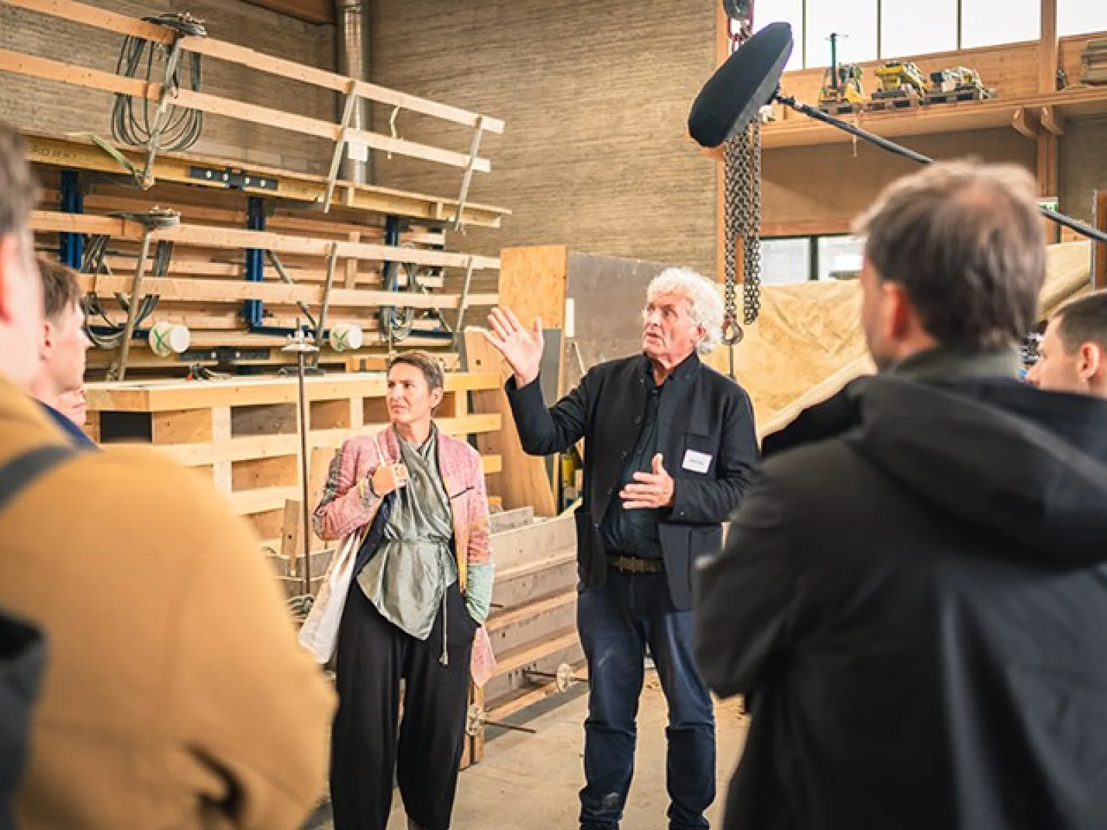 Eine Gruppe von Menschen steht in einer Werkstatt. Ein Mann spricht gestikulierend zu den Anwesenden. Im Hintergrund sind Holzregale und Werkzeuge zu sehen.