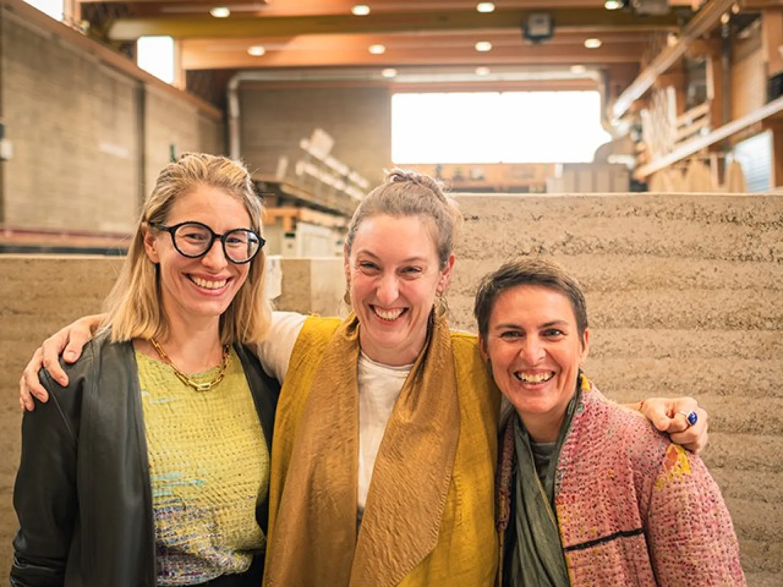 Drei lachende Personen stehen in einer Werkstatt vor Wänden aus Stampflehm. Sie tragen bunte Kleidung und umarmen sich freundschaftlich. Im Hintergrund sind Regale und Baumaterialien zu sehen.