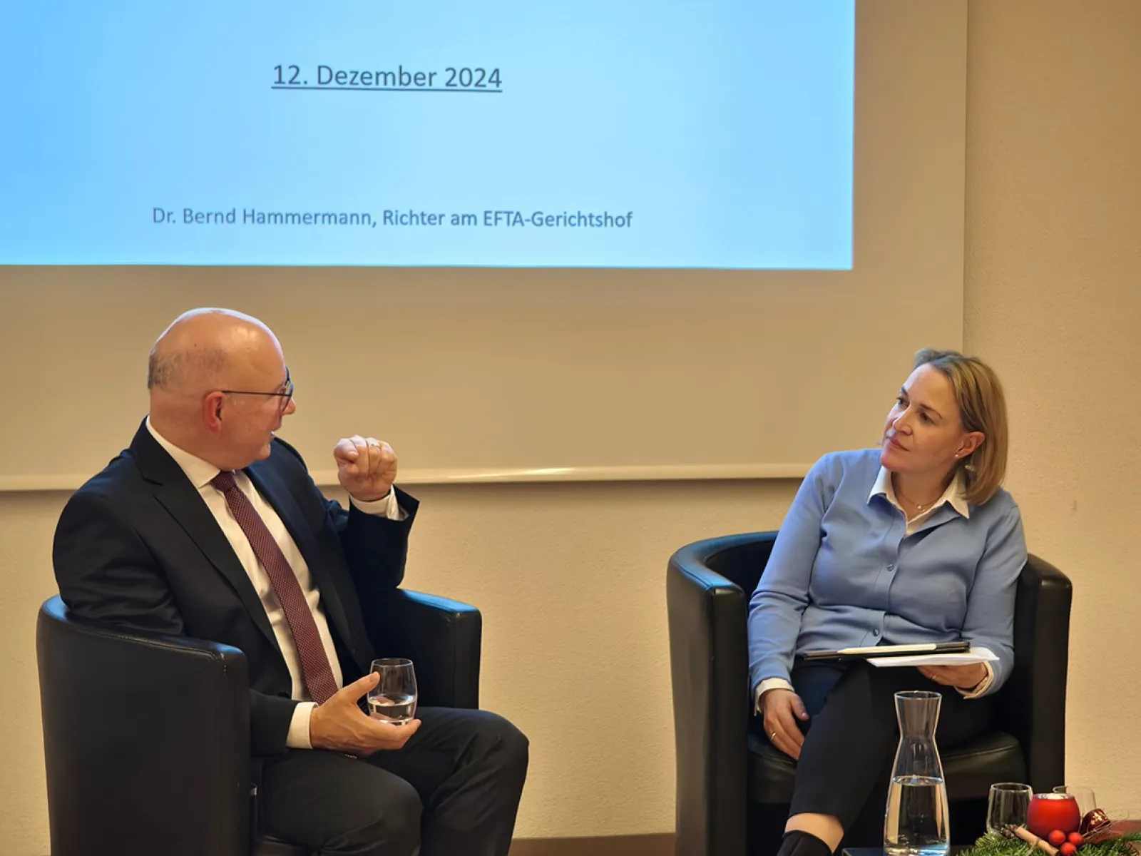 Zwei Personen sitzen in Sesseln vor einer Leinwand, die das Datum "12. Dezember 2024" zeigt. Eine Person hält ein Glas Wasser, die andere hat ein Notizbuch auf dem Schoß. Im Vordergrund steht eine Karaffe auf einem Tisch mit weihnachtlicher Dekoration.