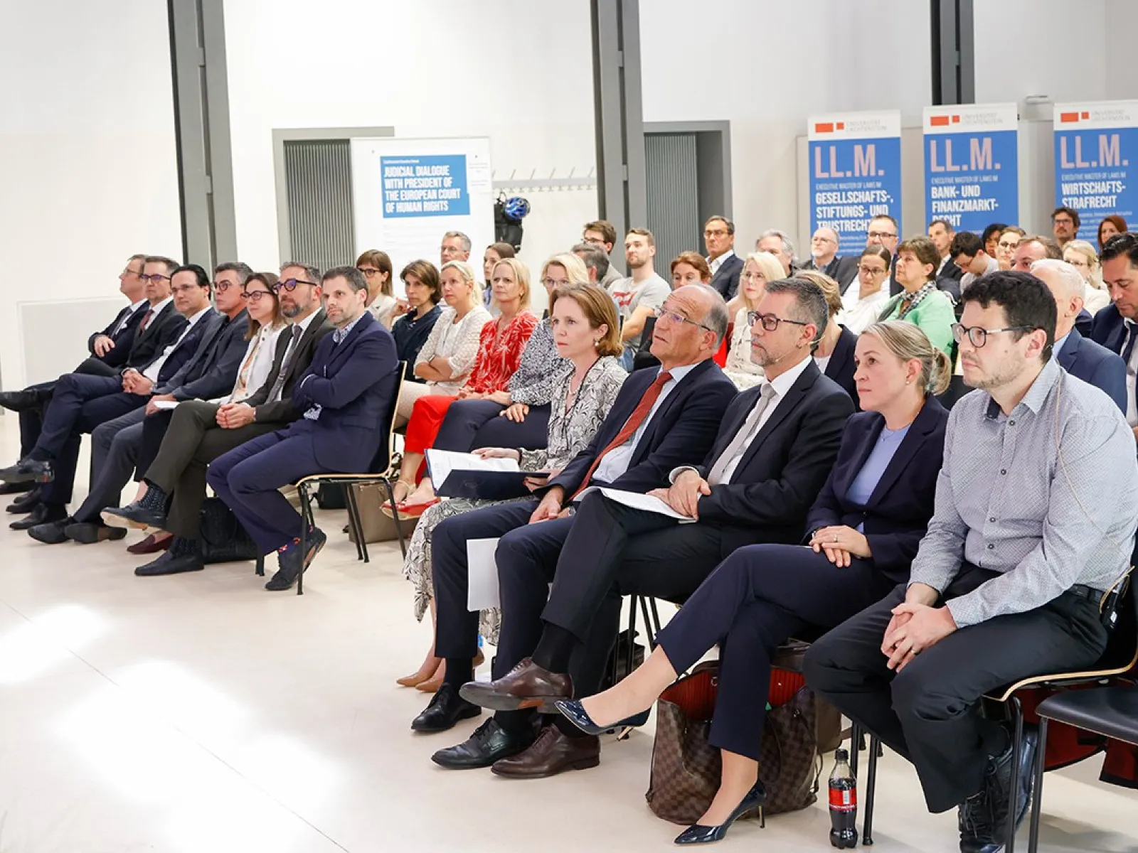 Eine Gruppe von Menschen sitzt in einem Konferenzraum der Universität Liechtenstein. Sie hören aufmerksam einem Vortrag zu. Im Hintergrund sind Banner mit der Aufschrift "LL.M." zu sehen.