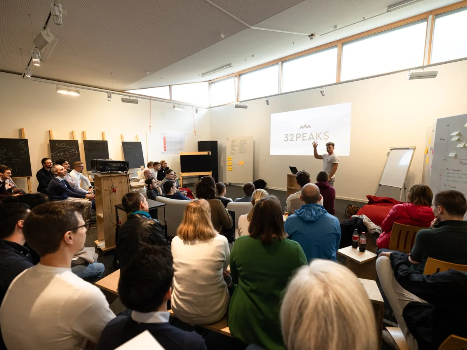 Eine Gruppe von Menschen sitzt in einem modernen Seminarraum und hört einem Vortragenden zu, der vor einer Leinwand mit der Aufschrift "32PEAKS" steht. Der Raum ist hell und mit großen Fenstern ausgestattet.