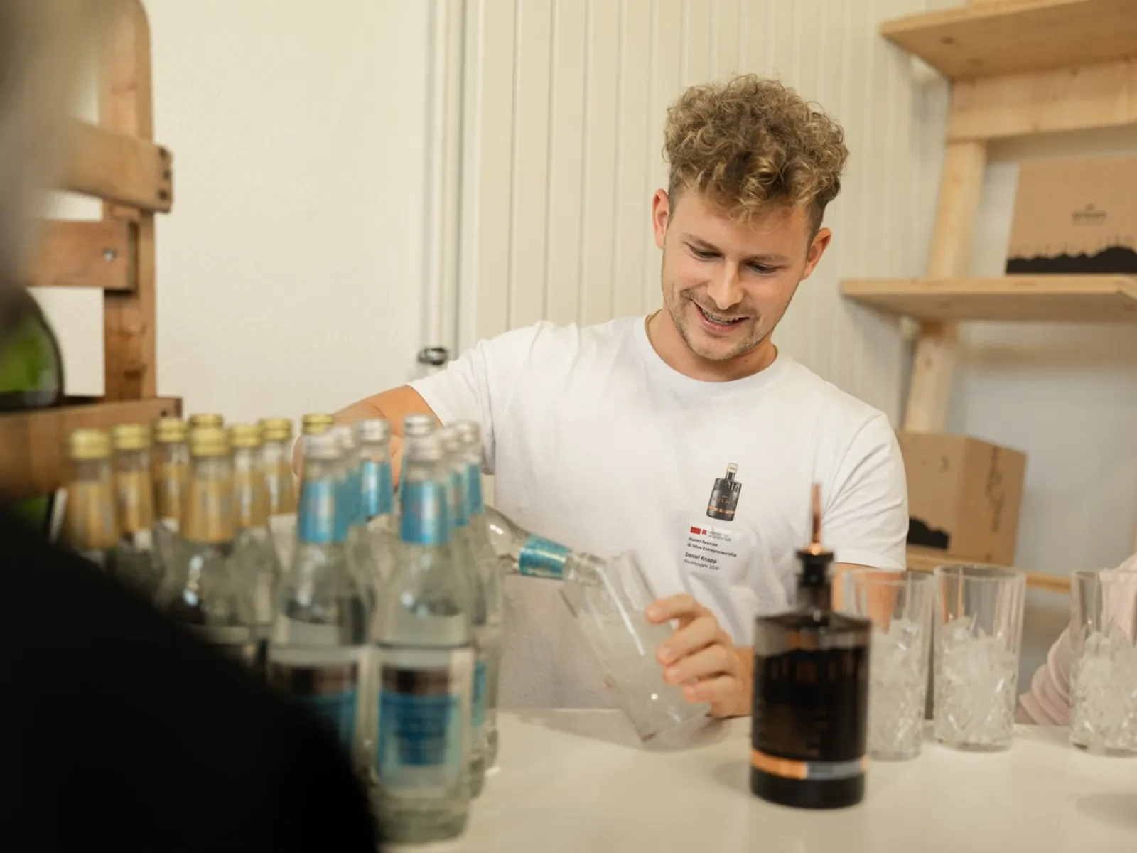 Ein junger Mann schenkt an einem Tisch Getränke ein. Auf dem Tisch stehen mehrere Flaschen und Gläser. Im Hintergrund sind Regale mit weiteren Gegenständen zu sehen.