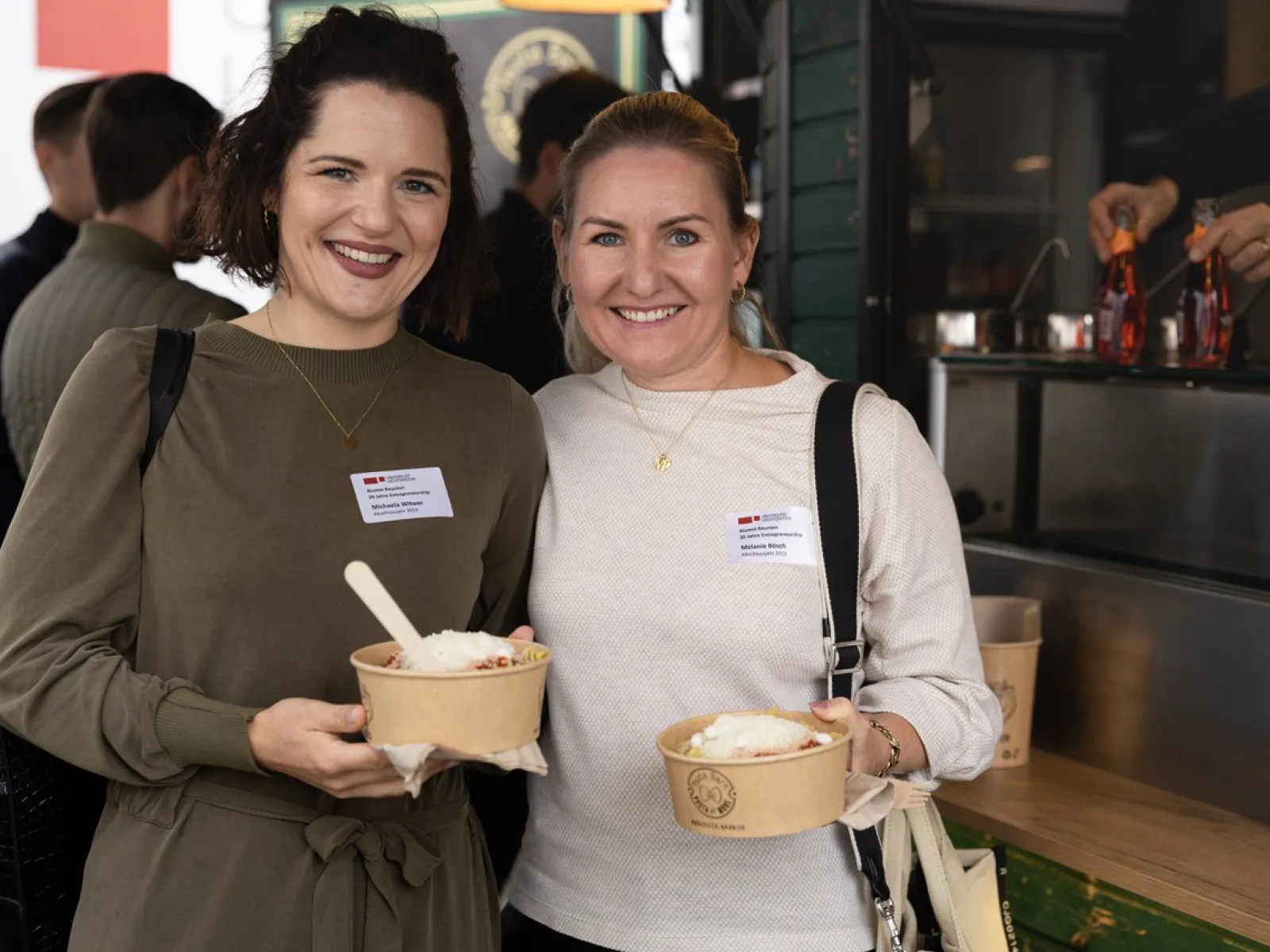 Zwei lächelnde Personen halten Schalen mit Essen in den Händen, während sie bei einer Feier an der Universität Liechtenstein stehen. Im Hintergrund ist ein Essensstand zu sehen.