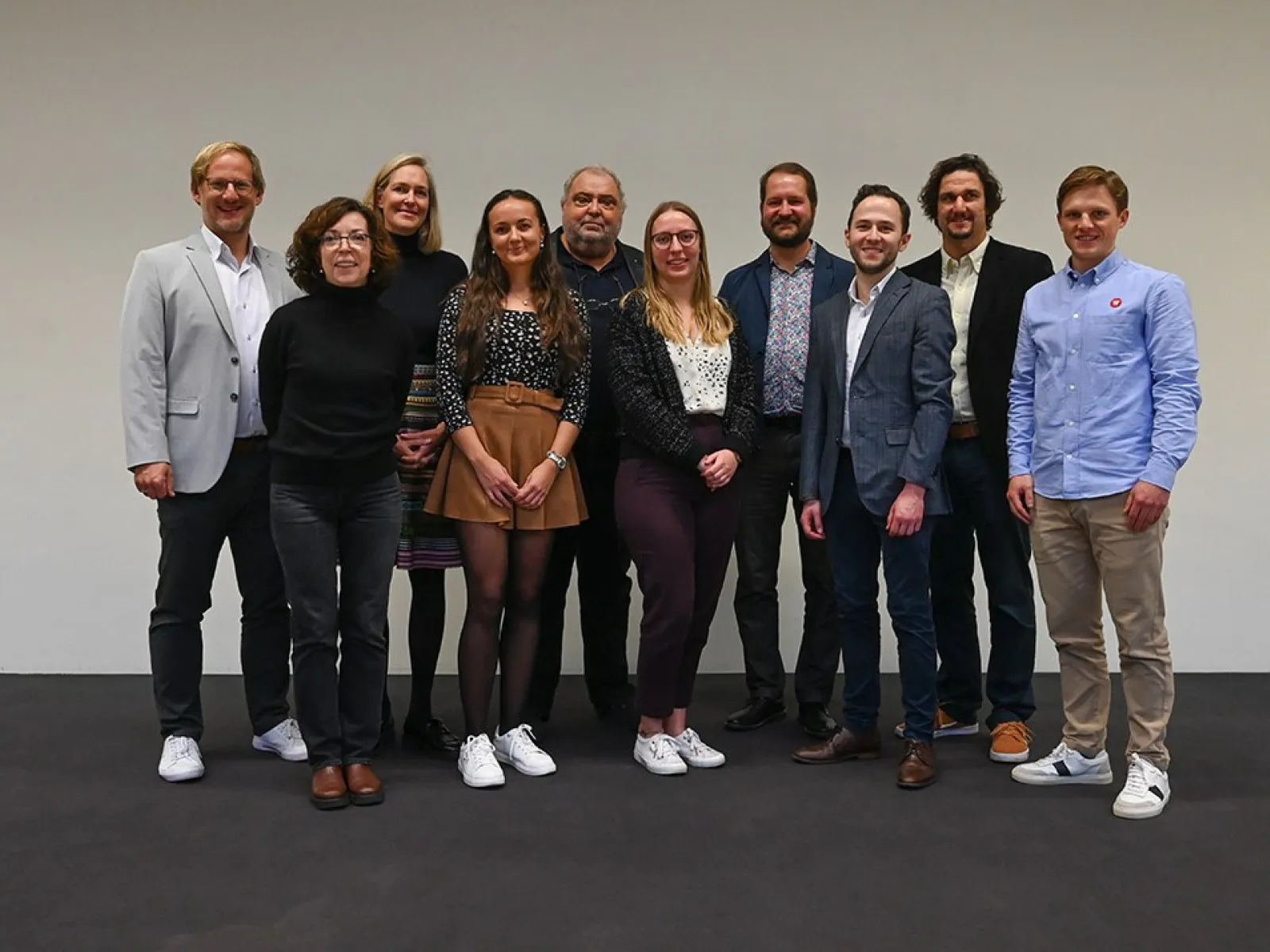Eine Gruppe von zehn Personen steht lächelnd nebeneinander vor einem neutralen Hintergrund. Sie tragen unterschiedliche, teils formelle, teils legere Kleidung. Das Bild vermittelt eine freundliche und professionelle Atmosphäre.