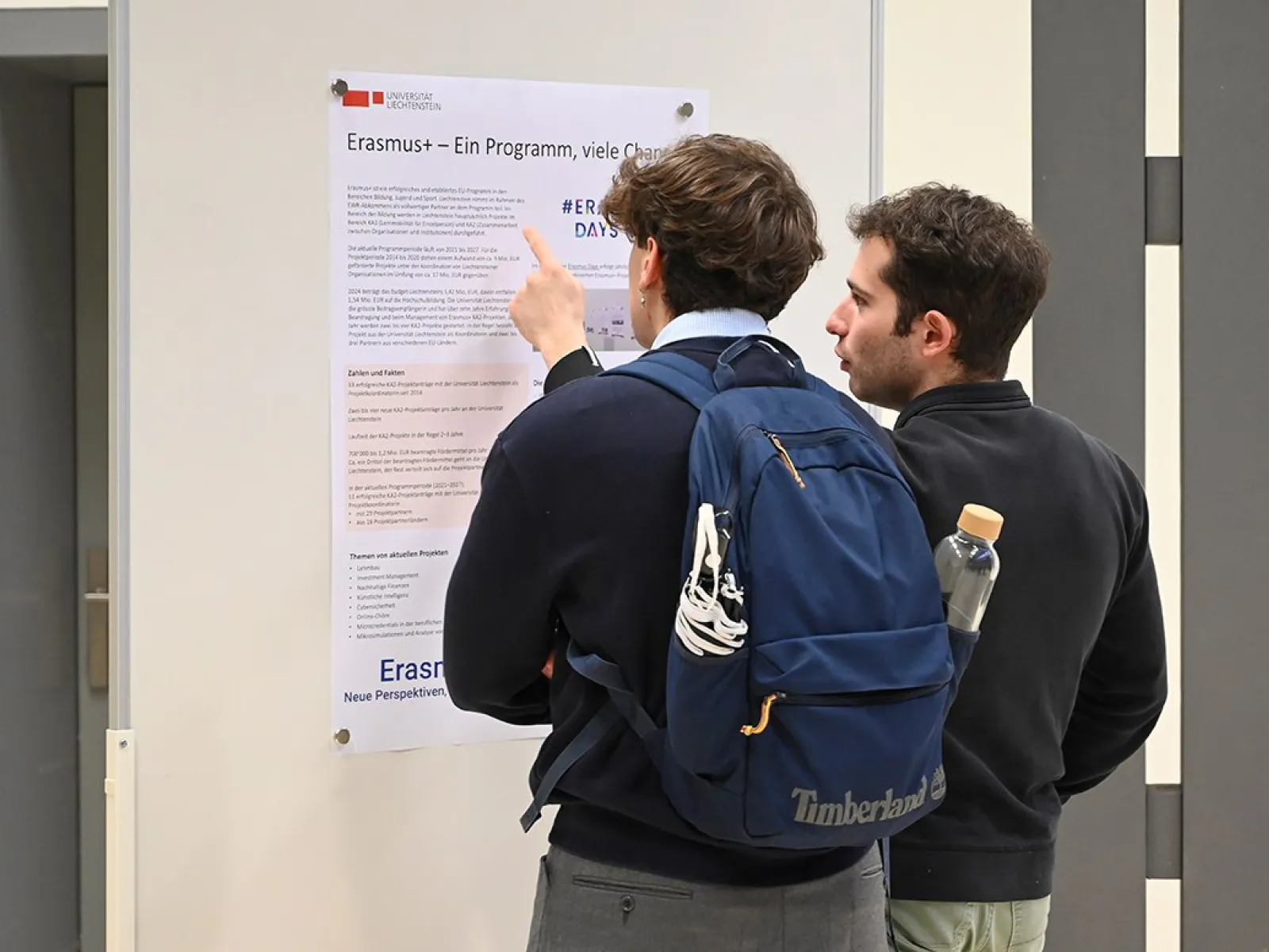 Zwei Personen betrachten ein Poster über das Erasmus-Programm an einer Wand. Eine Person zeigt auf den Text. Beide tragen Freizeitkleidung, einer hat einen Rucksack.