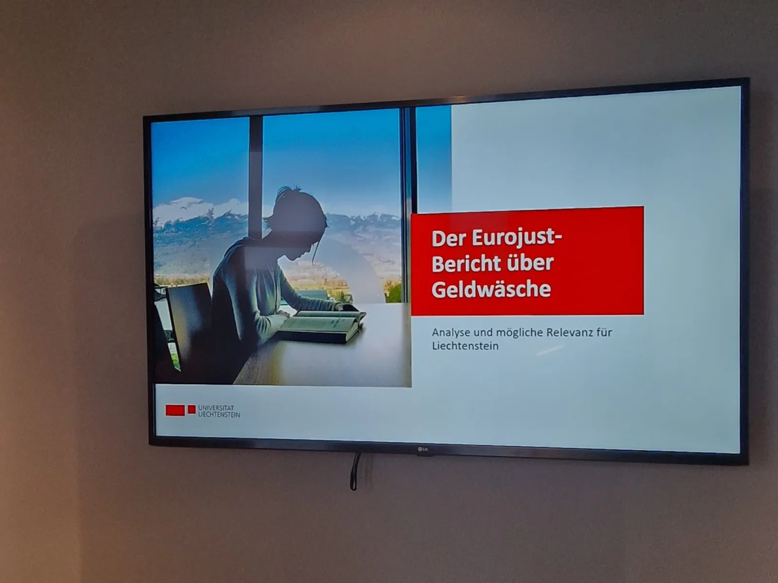Ein Bildschirm zeigt eine Präsentation mit dem Titel „Der Eurojust-Bericht über Geldwäsche: Analyse und mögliche Relevanz für Liechtenstein“. Links ist das Bild einer Person am Laptop vor einer Berglandschaft zu sehen.