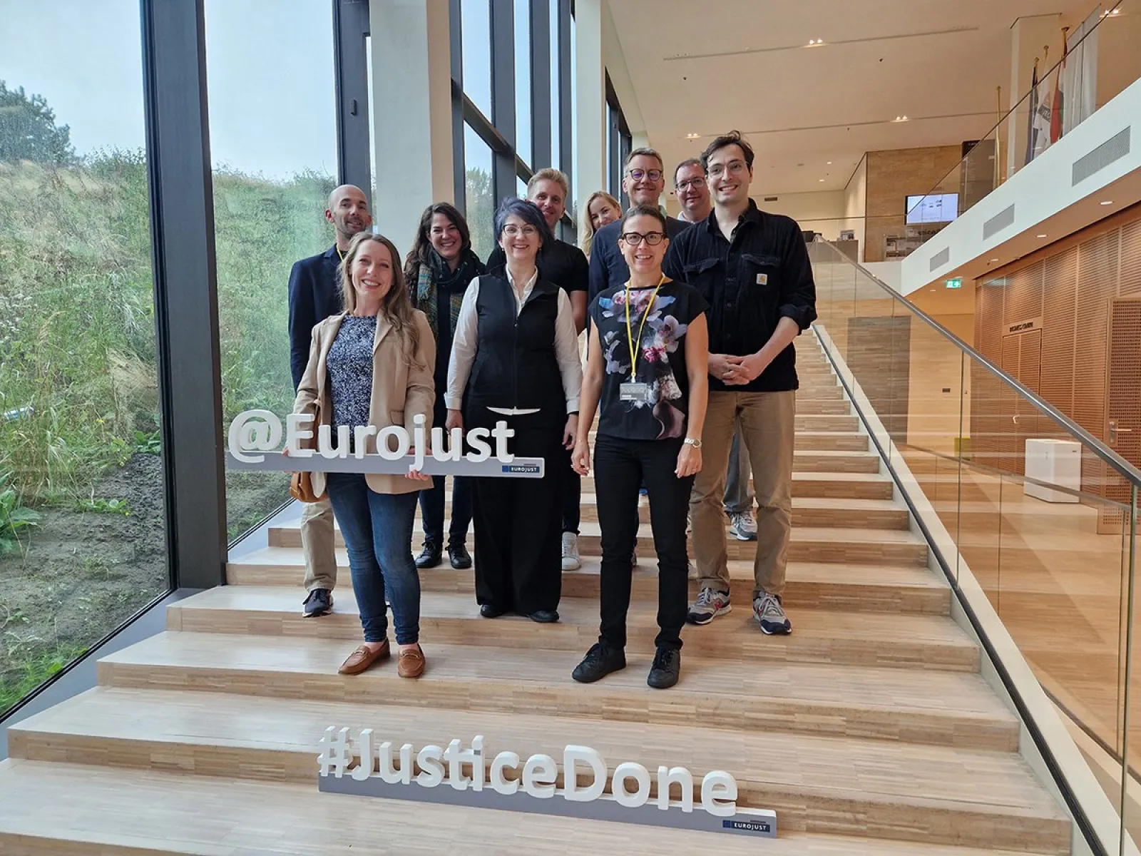 Gruppe von Studierenden und Dozenten des Executive Master of Laws (LL.M.) im Wirtschaftsstrafrecht posiert auf einer Treppe bei Eurojust in Den Haag. Im Vordergrund sind die Schriftzüge "@Eurojust" und "#JusticeDone" zu sehen.