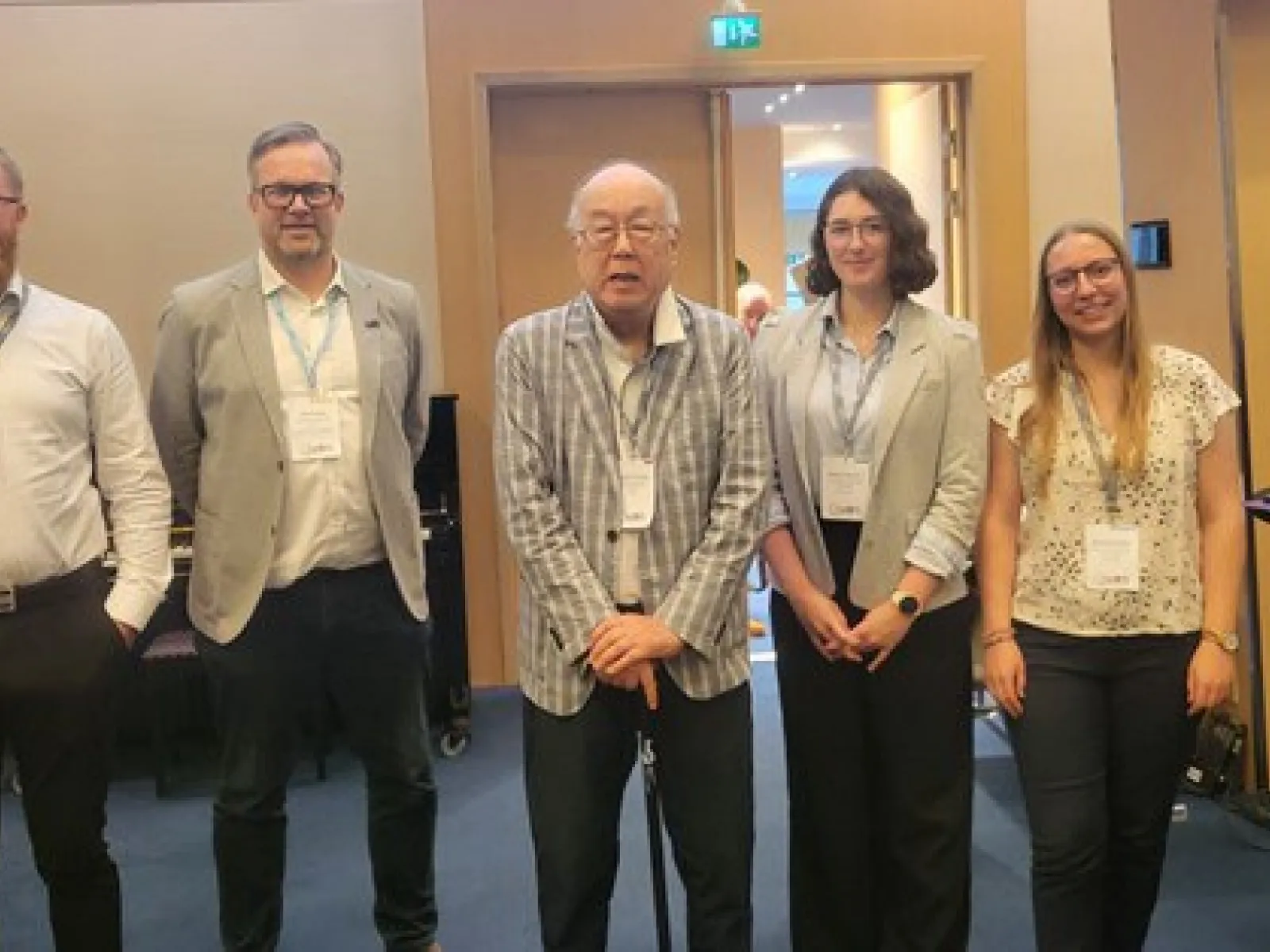 Fünf Personen stehen nebeneinander in einem Konferenzraum. Sie tragen Namensschilder und lächeln in die Kamera. Im Hintergrund sind Türen und ein Tisch sichtbar.