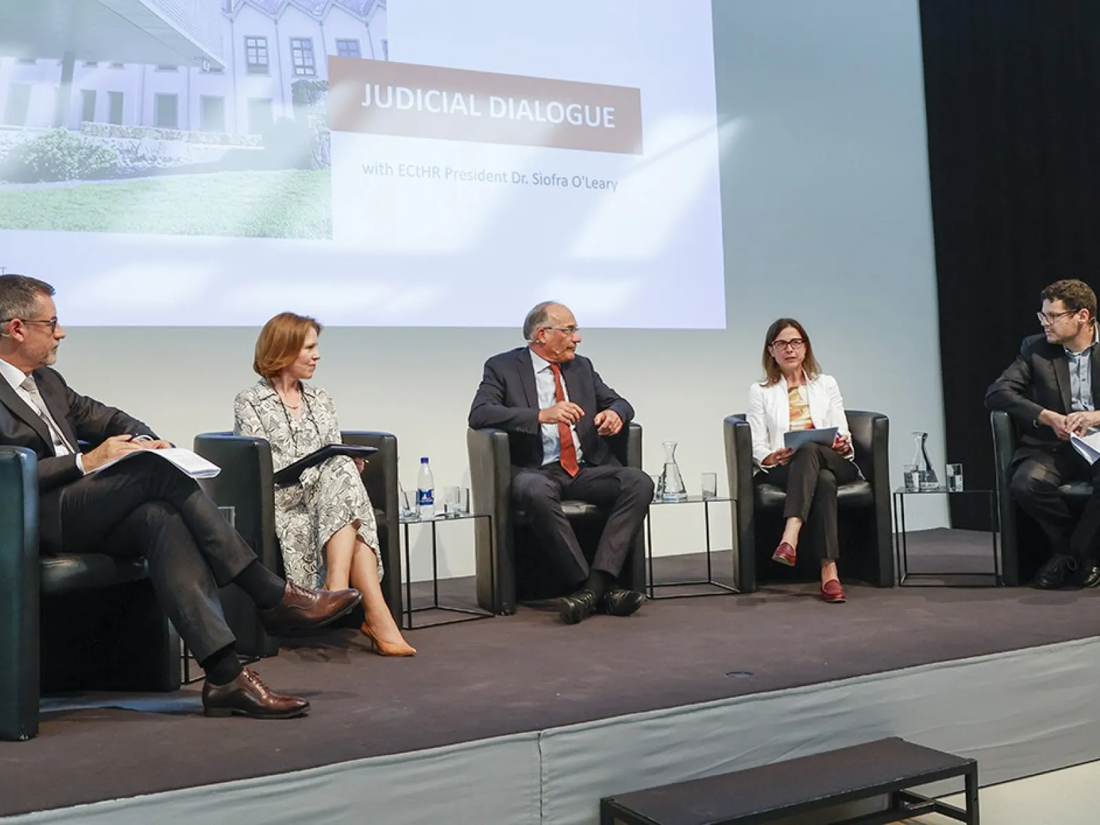 Podiumsdiskussion an der Universität Liechtenstein mit fünf Personen auf einer Bühne, die an einem juristischen Dialog teilnehmen. Im Hintergrund ist eine Präsentation mit dem Titel "Judicial Dialogue" zu sehen.