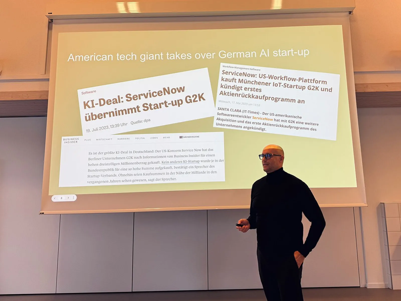 Ein Vortragender steht vor einer Präsentationsfolie, die Schlagzeilen über die Übernahme des deutschen KI-Start-ups G2K durch den amerikanischen Technologiekonzern ServiceNow zeigt.
