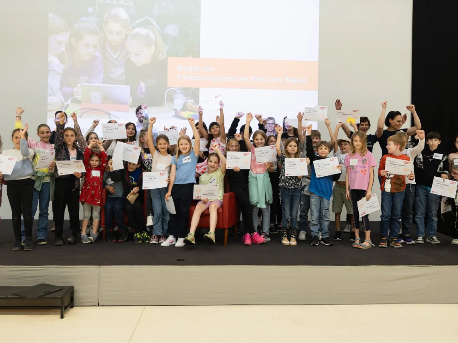 Gruppe von Kindern auf einer Bühne, die stolz Zertifikate hochhalten, im Rahmen der Kinder-Uni an der Universität Liechtenstein. Im Hintergrund ein Bildschirm mit der Aufschrift „Entdeckungsreise ins Reich der Regeln“.