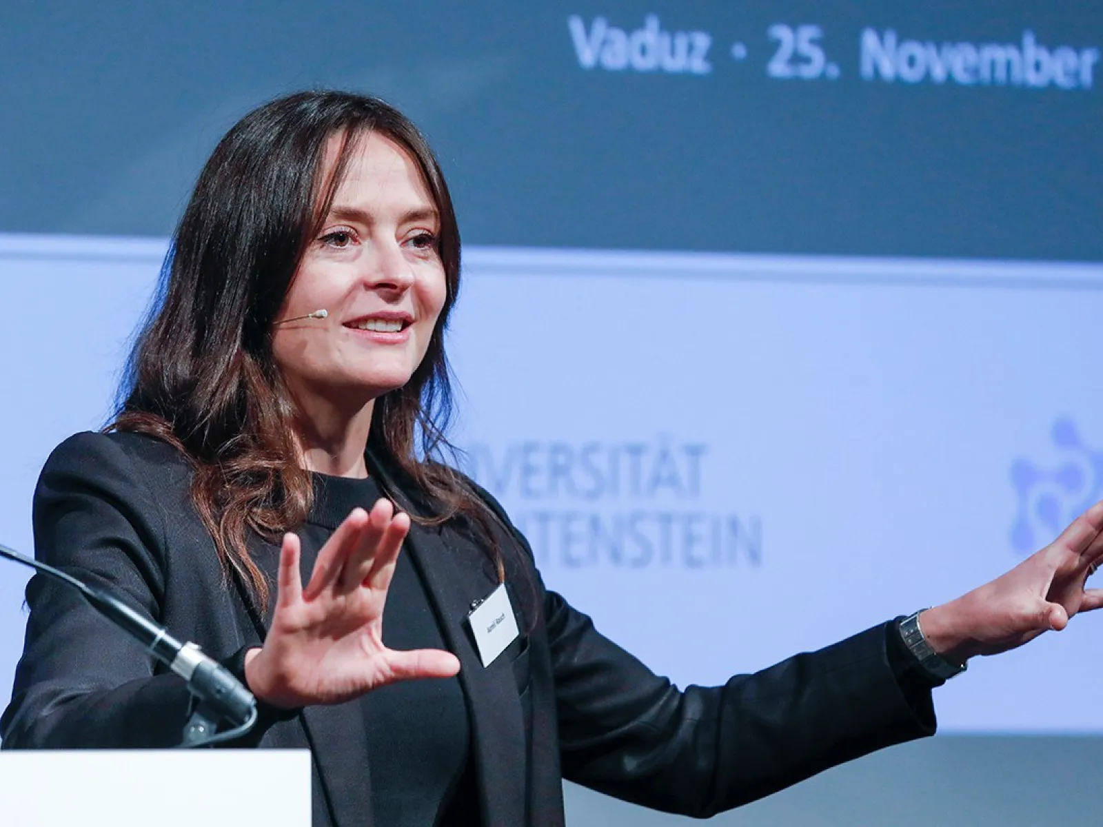 Eine Frau hält einen Vortrag bei einer Konferenz an der Universität Liechtenstein. Sie trägt ein Mikrofon und gestikuliert mit den Händen. Im Hintergrund ist ein Banner mit dem Datum der Veranstaltung und dem Logo der Universität zu sehen.
