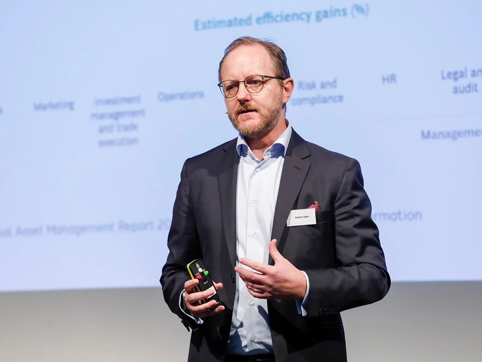 Ein Redner auf einer Konferenz präsentiert vor einer Leinwand mit Diagrammen und Texten. Er trägt einen Anzug und hält einen Präsentationsklicker in der Hand.