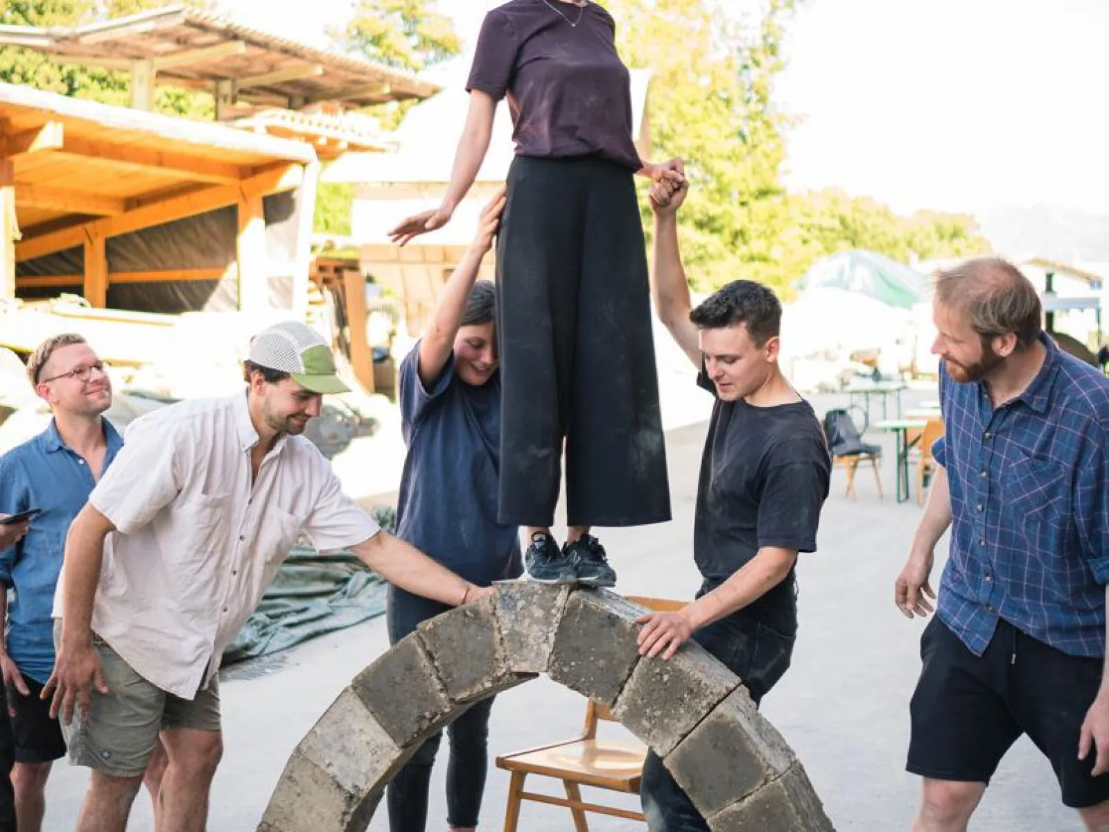 Ein Gruppe von Menschen baut einen selbsttragenden Lehmbogen im Freien. Eine Person steht oben auf dem Bogen, während andere sie stützen. Im Hintergrund sind Bäume und Gebäude zu sehen.