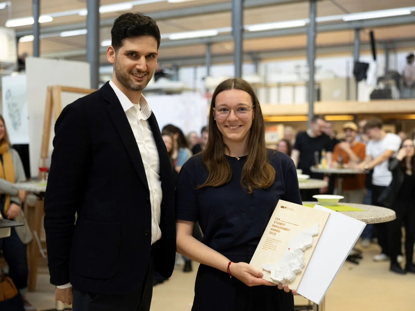Zwei Personen stehen in einem Innenraum. Eine Person hält eine Auszeichnung oder ein Buch in den Händen. Im Hintergrund sind weitere Menschen und eine helle, moderne Umgebung zu sehen.