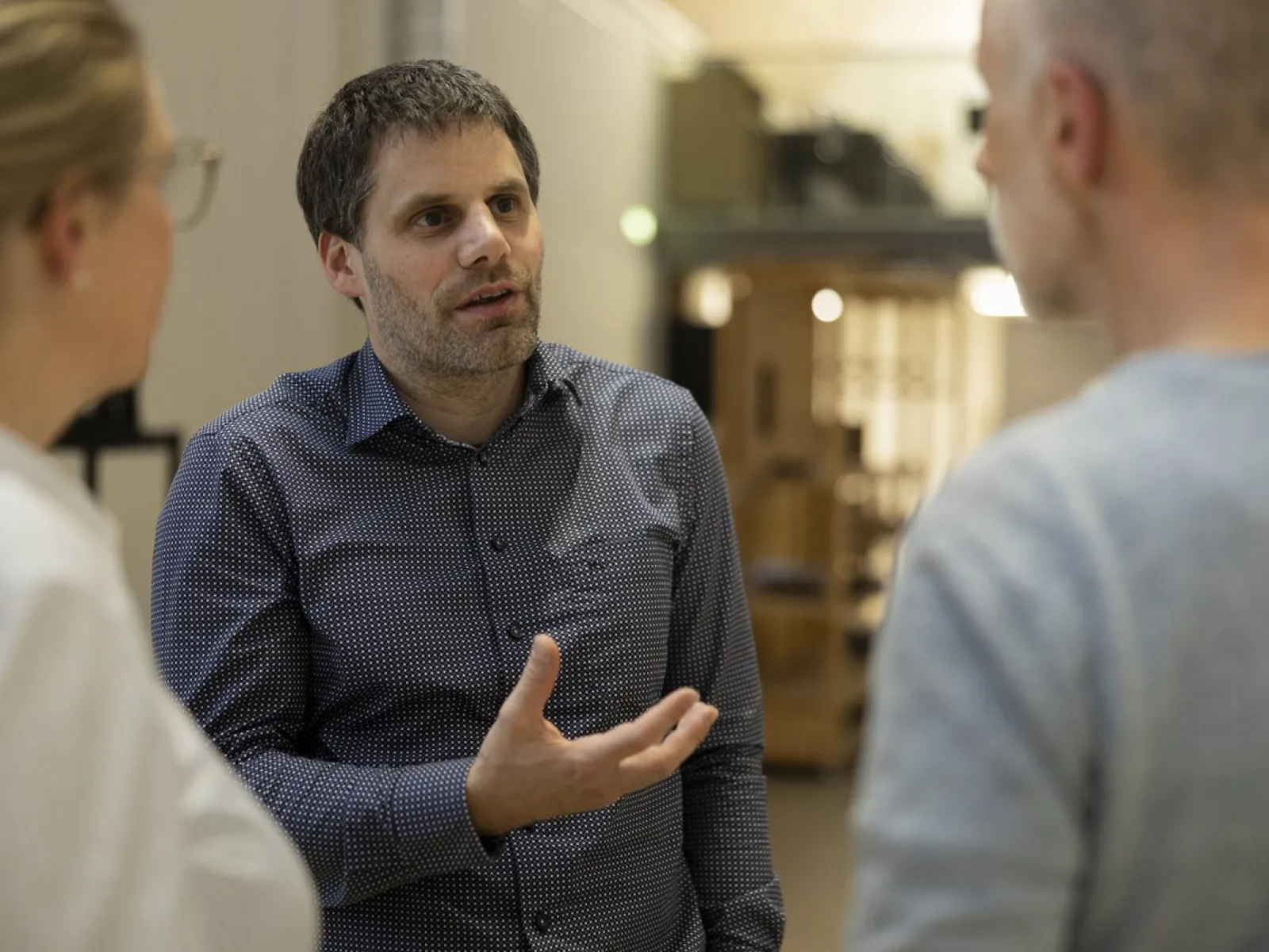 Ein Mann in einem karierten Hemd spricht gestikulierend mit zwei anderen Personen in einem Innenraum. Die Szene wirkt professionell und konzentriert.