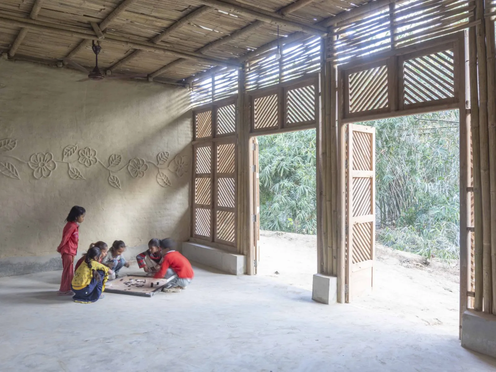 Einige Kinder spielen auf dem Boden eines hellen, nachhaltig gebauten Raumes mit großen Holztüren und dekorativen Wänden.